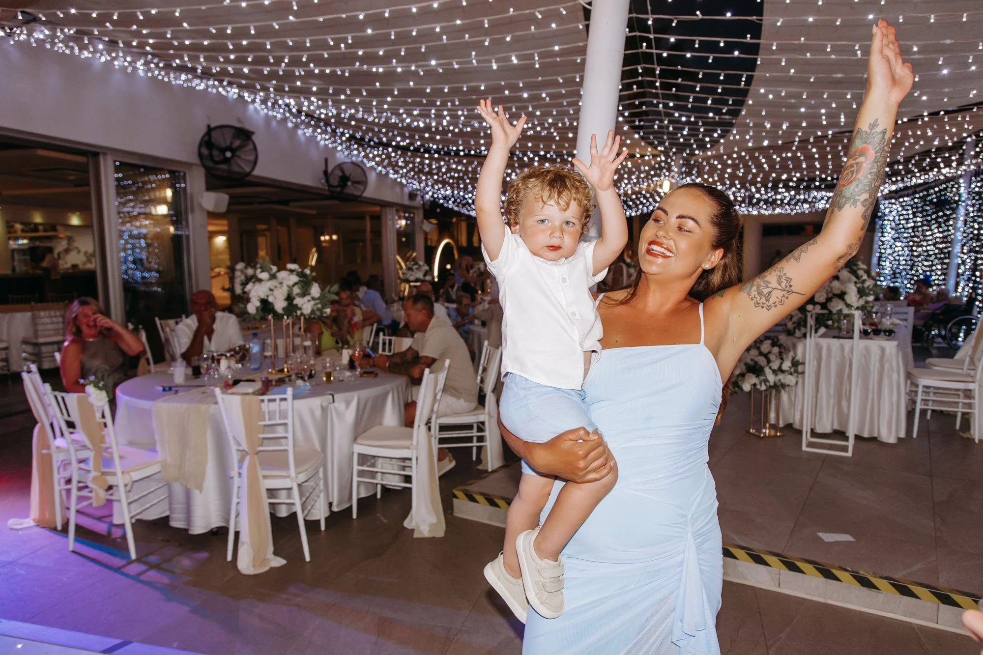 A woman in a light blue dress holding a young boy in a white shirt and light blue shorts, both with their hands raised, celebrating at a decorated indoor event with string lights, arranged tables, and guests in the background.