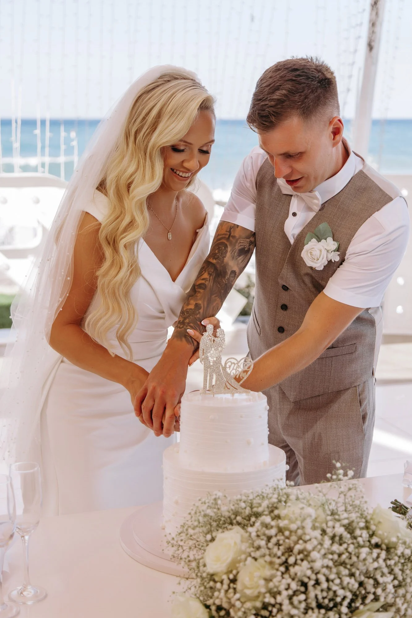 A bride and groom cut a wedding cake under a canopy by the seaside, with the ocean visible in the background.