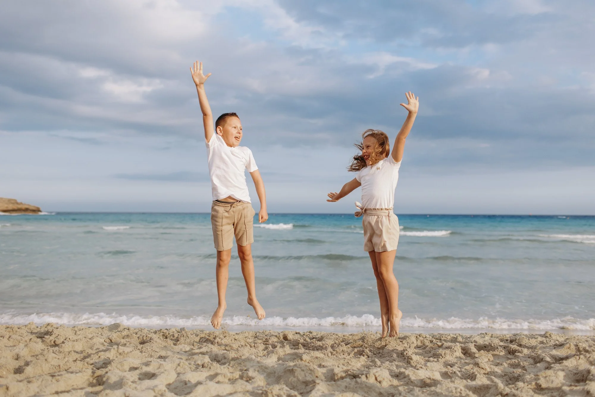 Two children, a boy and a girl, jumping together on a sandy beach with the ocean and a cloudy sky in the background.
