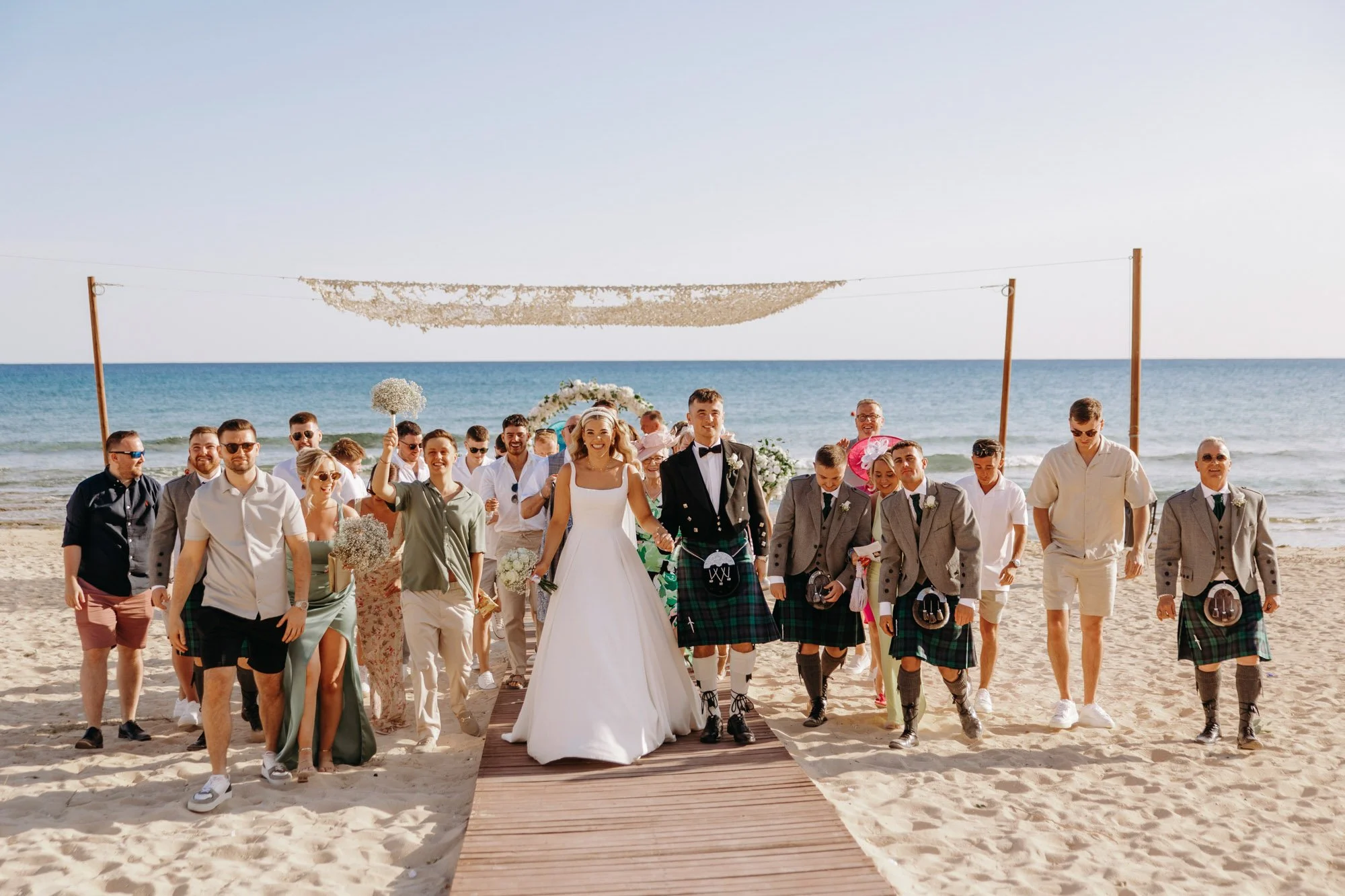 A wedding party walking on the beach, with the bride and groom in front, all smiling and holding hands. The bride is wearing a white wedding dress and the groom is in a traditional Scottish kilt. The sky is clear, and the ocean is in the background.