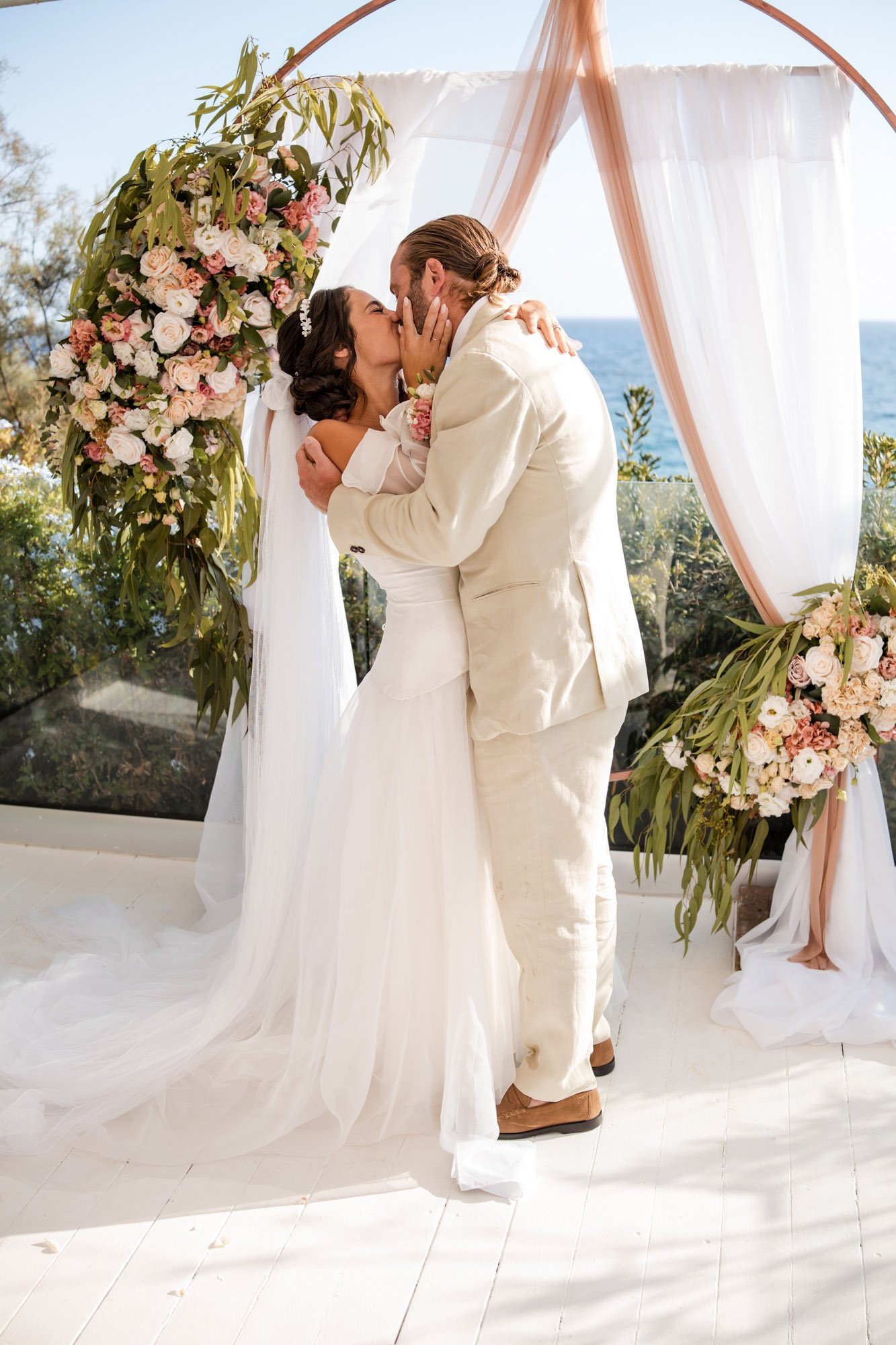A bride and groom share a kiss during their outdoor wedding ceremony. The bride wears a white gown and veil, and the groom wears a cream suit. They stand in front of a floral arch with pink and white flowers, and the ocean is visible in the backgroun