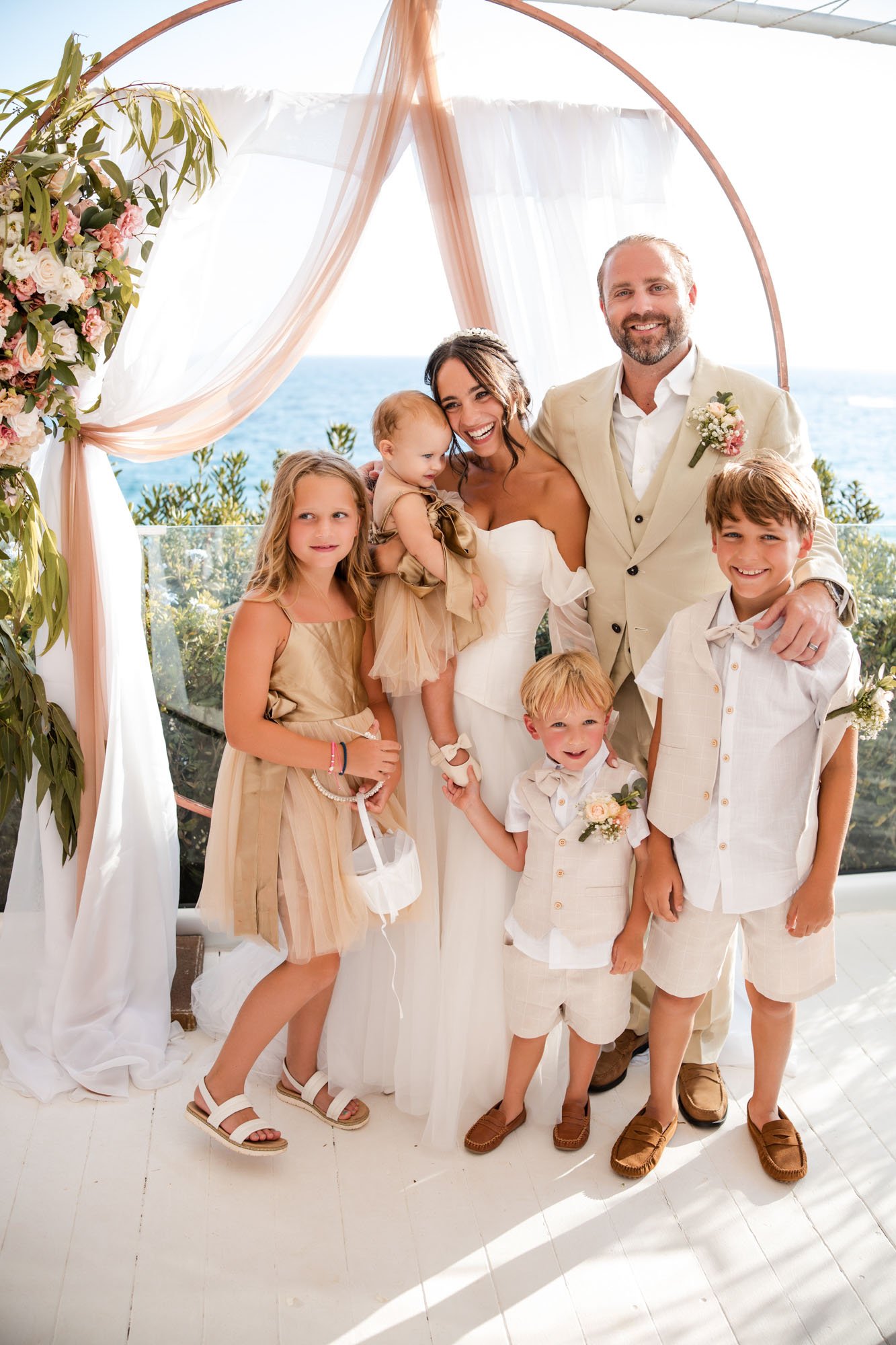 A family celebrating a wedding outdoors near the ocean, standing under a decorated arch with pink and white drapes and flowers.