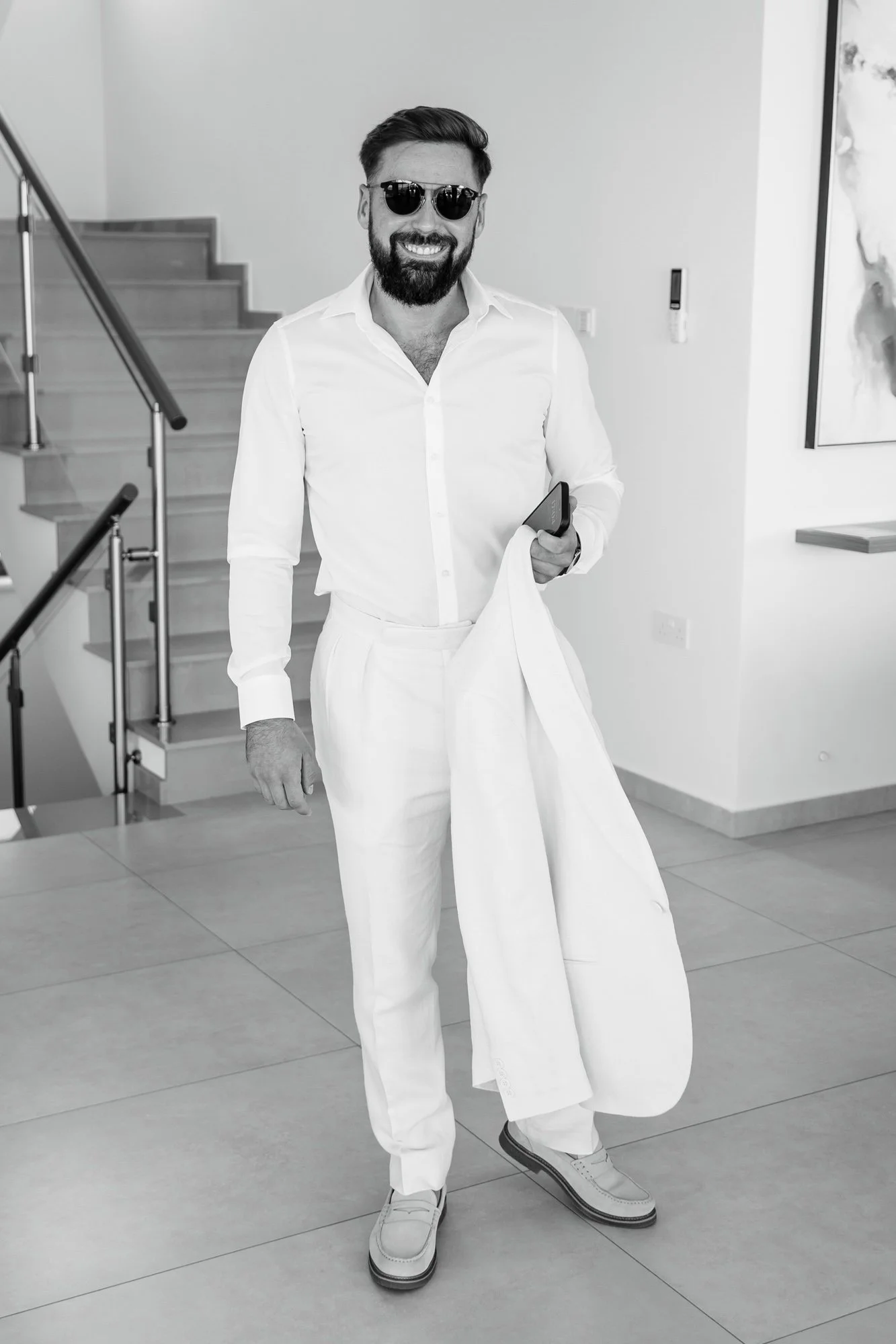 Man wearing dark sunglasses, white dress shirt, white pants, and white shoes, holding a jacket and a phone, standing indoors near stairs.