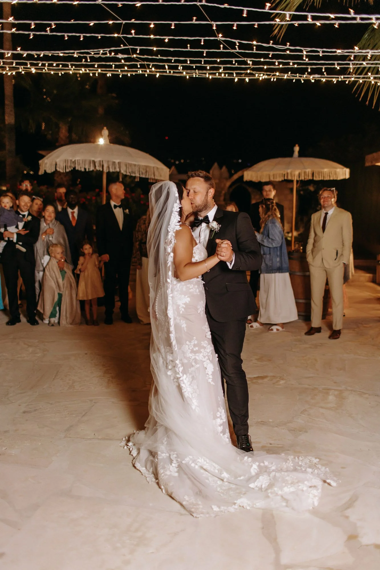 Bride and groom sharing a kiss during their wedding dance under string lights at night, with guests watching in the background.
