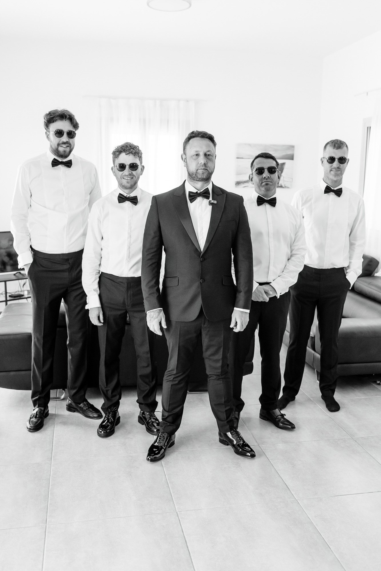 Group of six men dressed in tuxedos and bow ties, standing in a living room with a sofa, TV, and artwork on the wall, all wearing sunglasses.