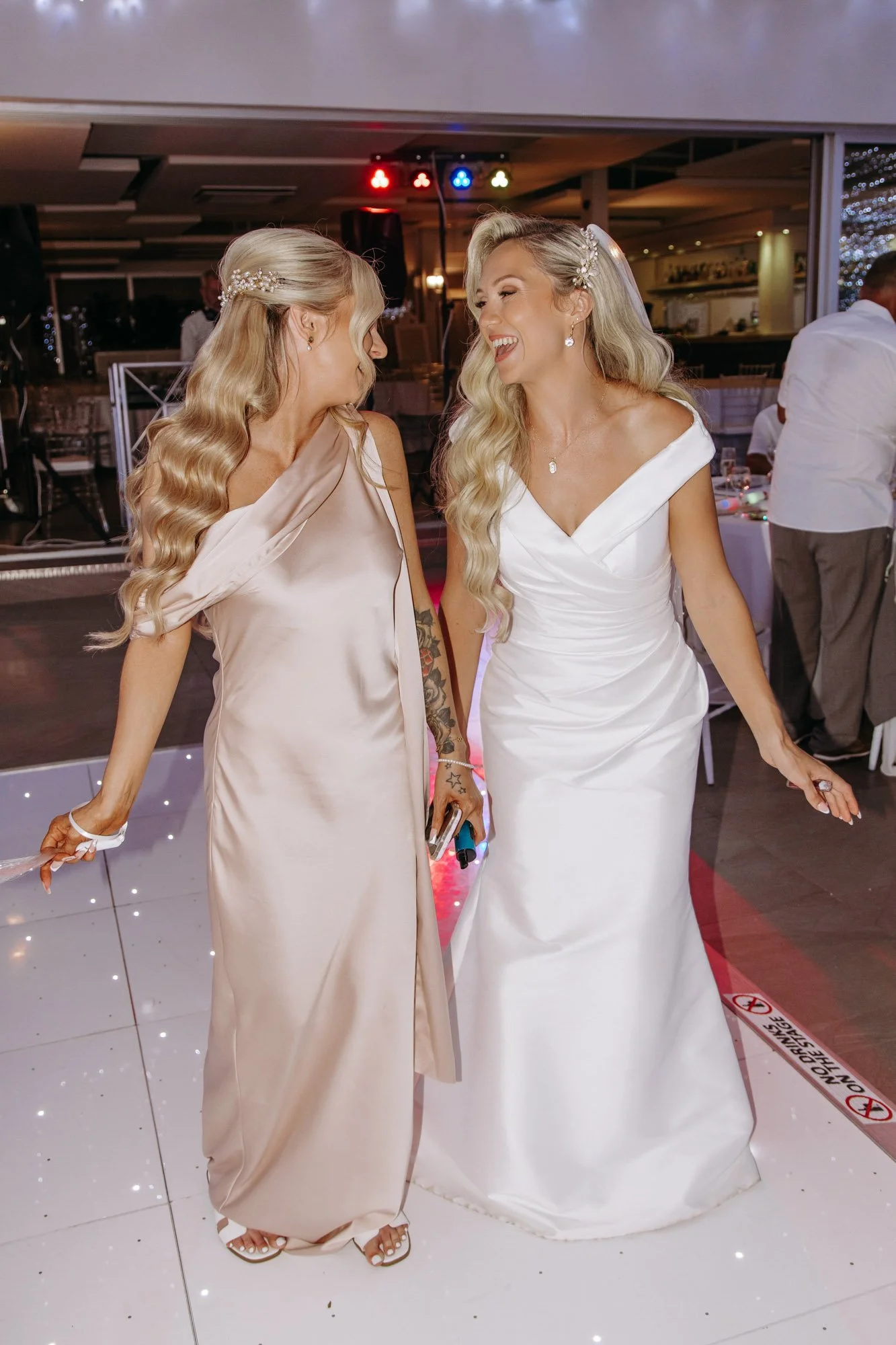 Two women, one in a white wedding dress and the other in a champagne-colored dress, are sharing a joyful moment at a wedding reception.