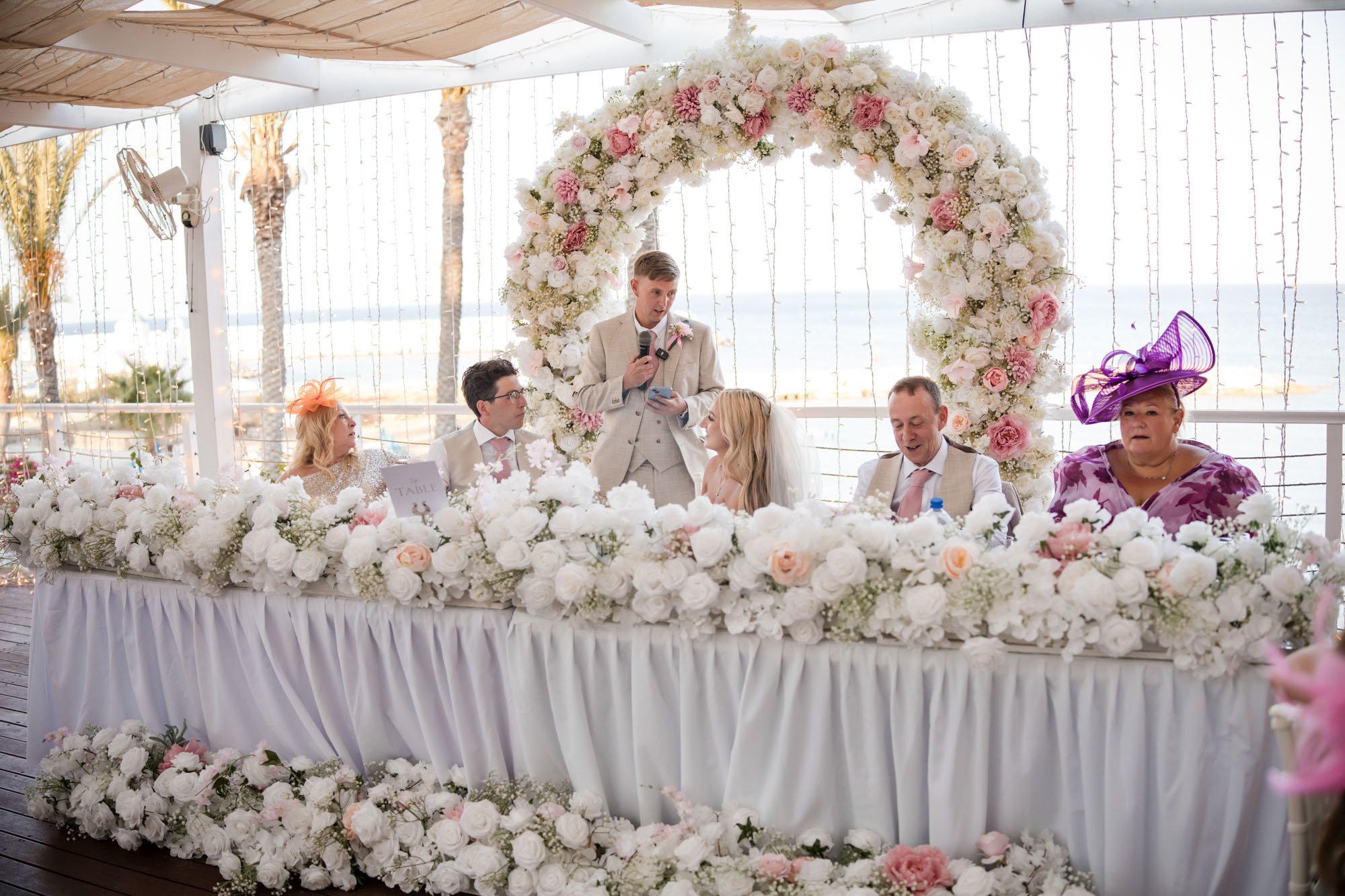 Wedding reception with six people seated at a table decorated with white and pink flowers, behind a floral arch, in an outdoor setting near the beach.