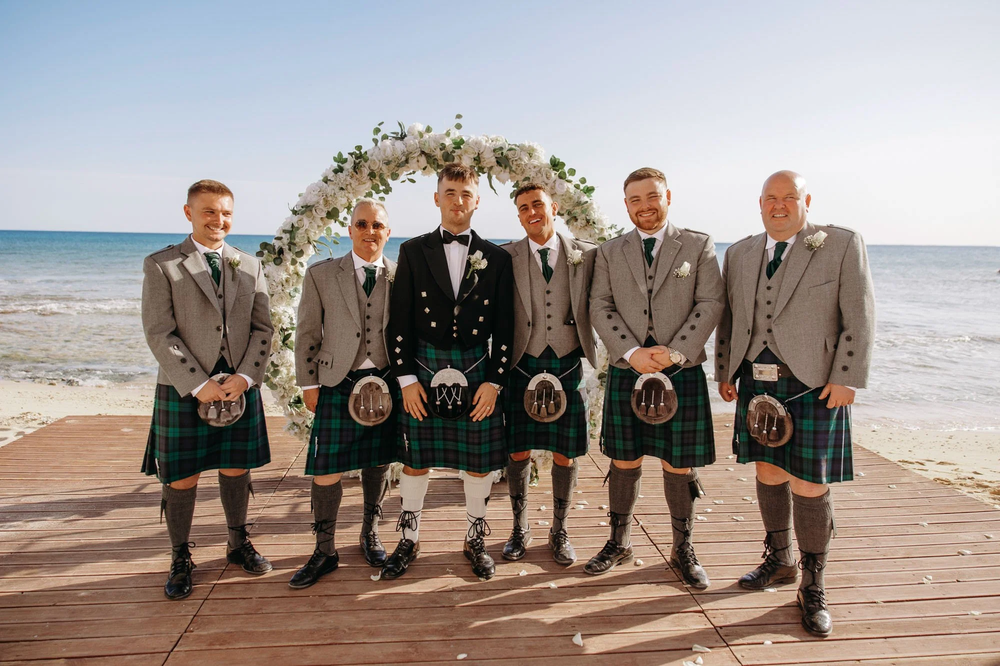 A group of seven men in kilts and jackets standing on a wooden platform on a beach during a wedding ceremony, with a floral arch in the background and the ocean behind them.