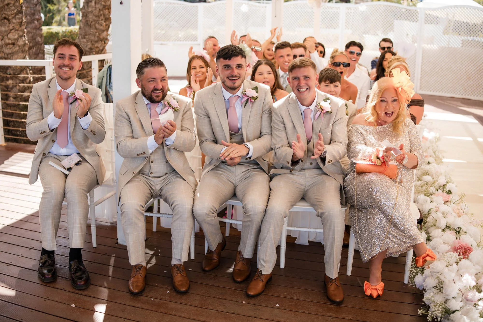 Group of wedding guests, mostly men in matching beige suits and pink ties, sitting and clapping during a wedding ceremony