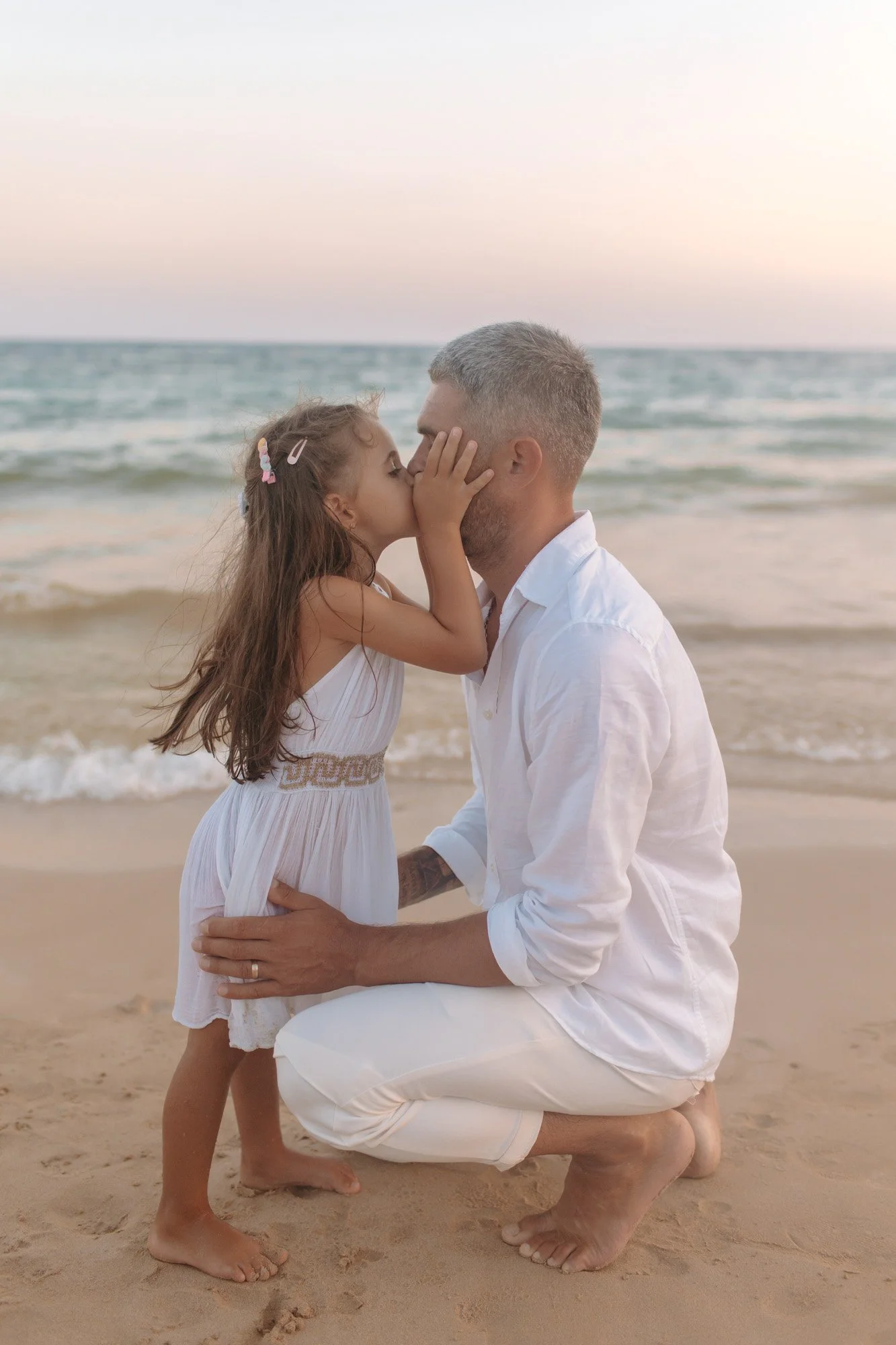 A man kneeling on the sand at the beach, holding a young girl, who is giving him a kiss on the nose. The scene is set during sunset, with gentle waves in the background.