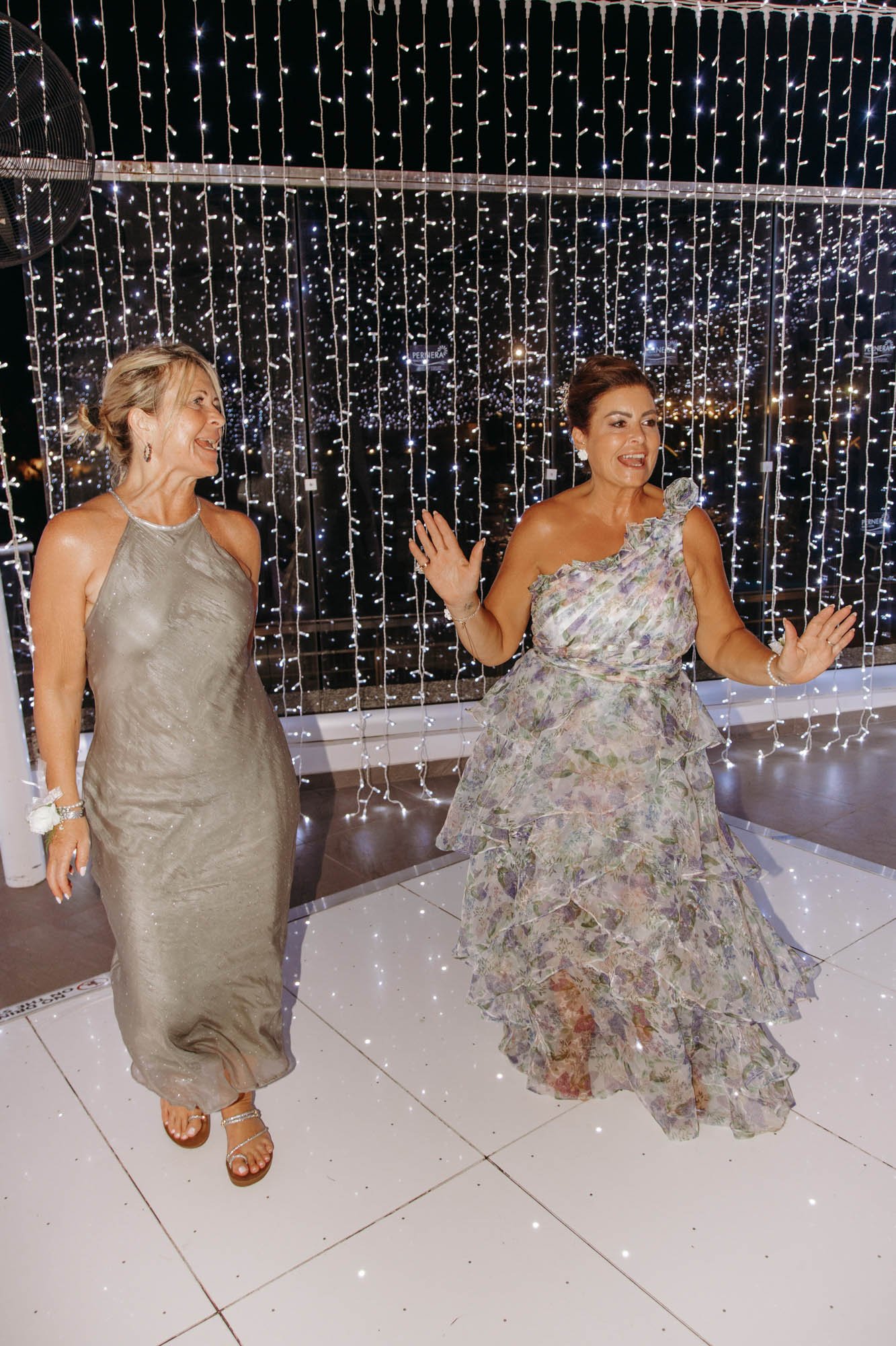 Two women dancing and smiling at a party or celebration with string lights hanging in the background.