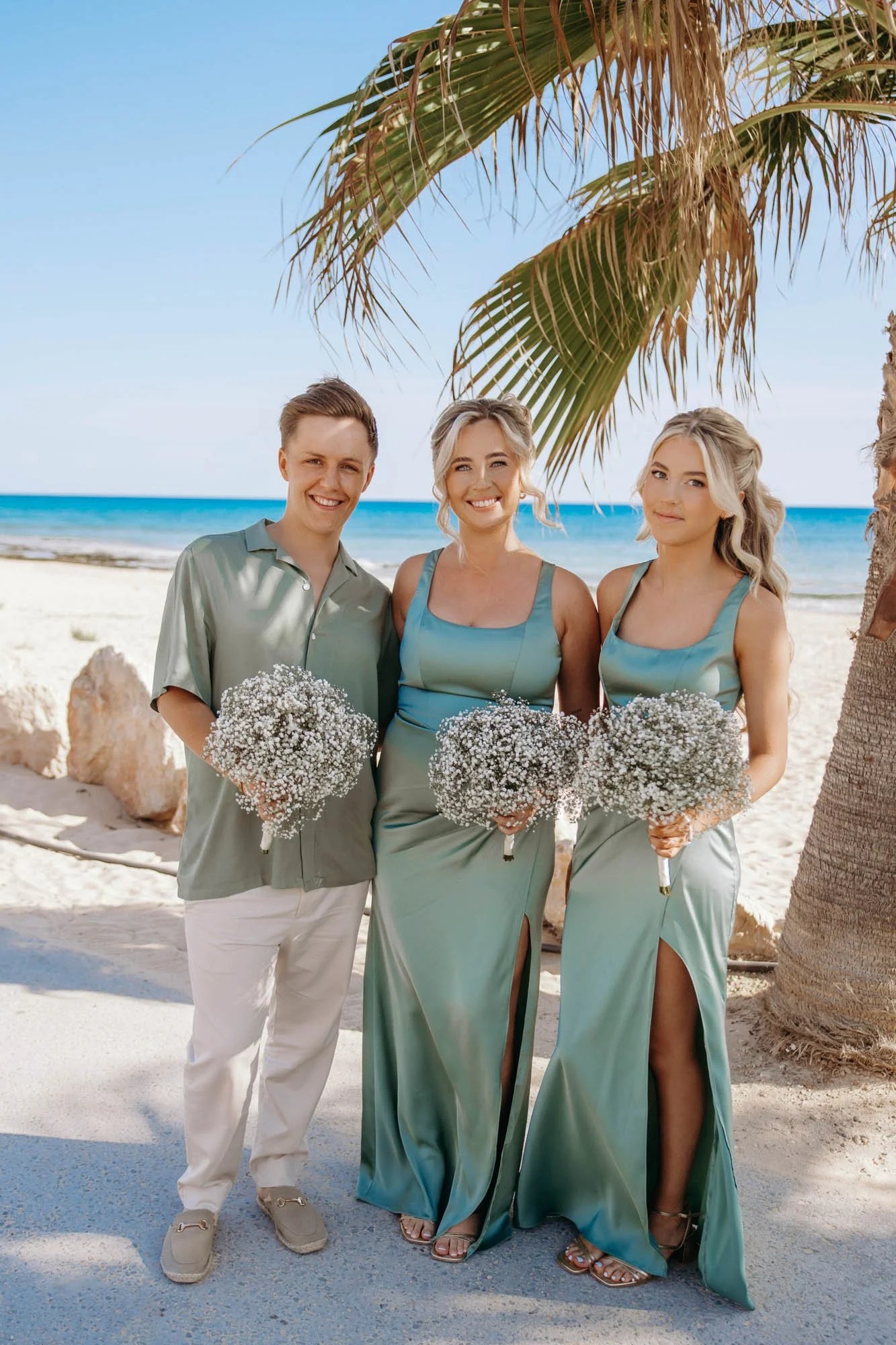 People standing on a beach, holding white flower bouquets, under a palm tree, with ocean waves in the background.