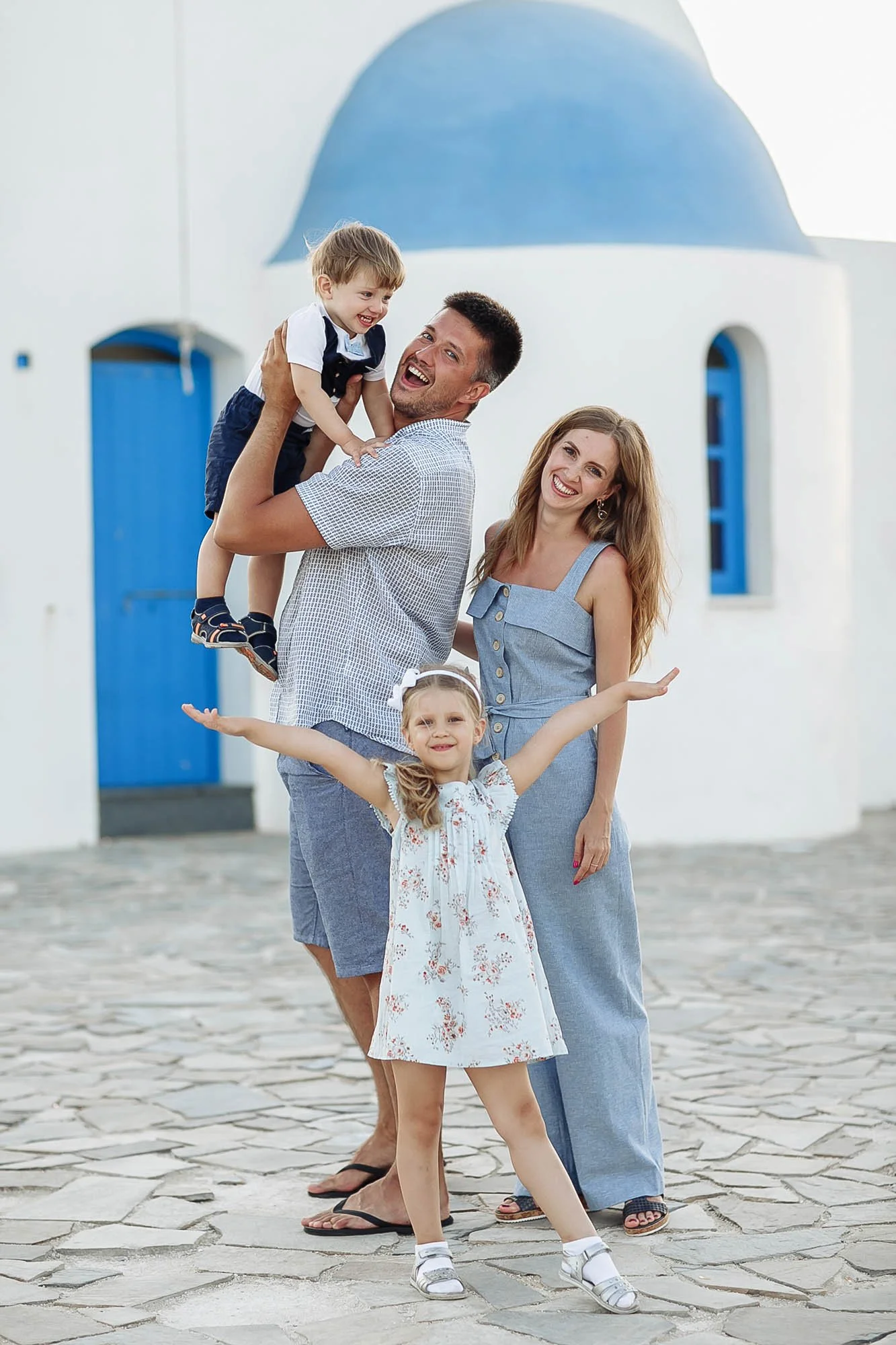 A family of four outdoors, with a white church with blue details in the background. The man is holding a young boy, and a woman and girl are standing nearby smiling and posing.