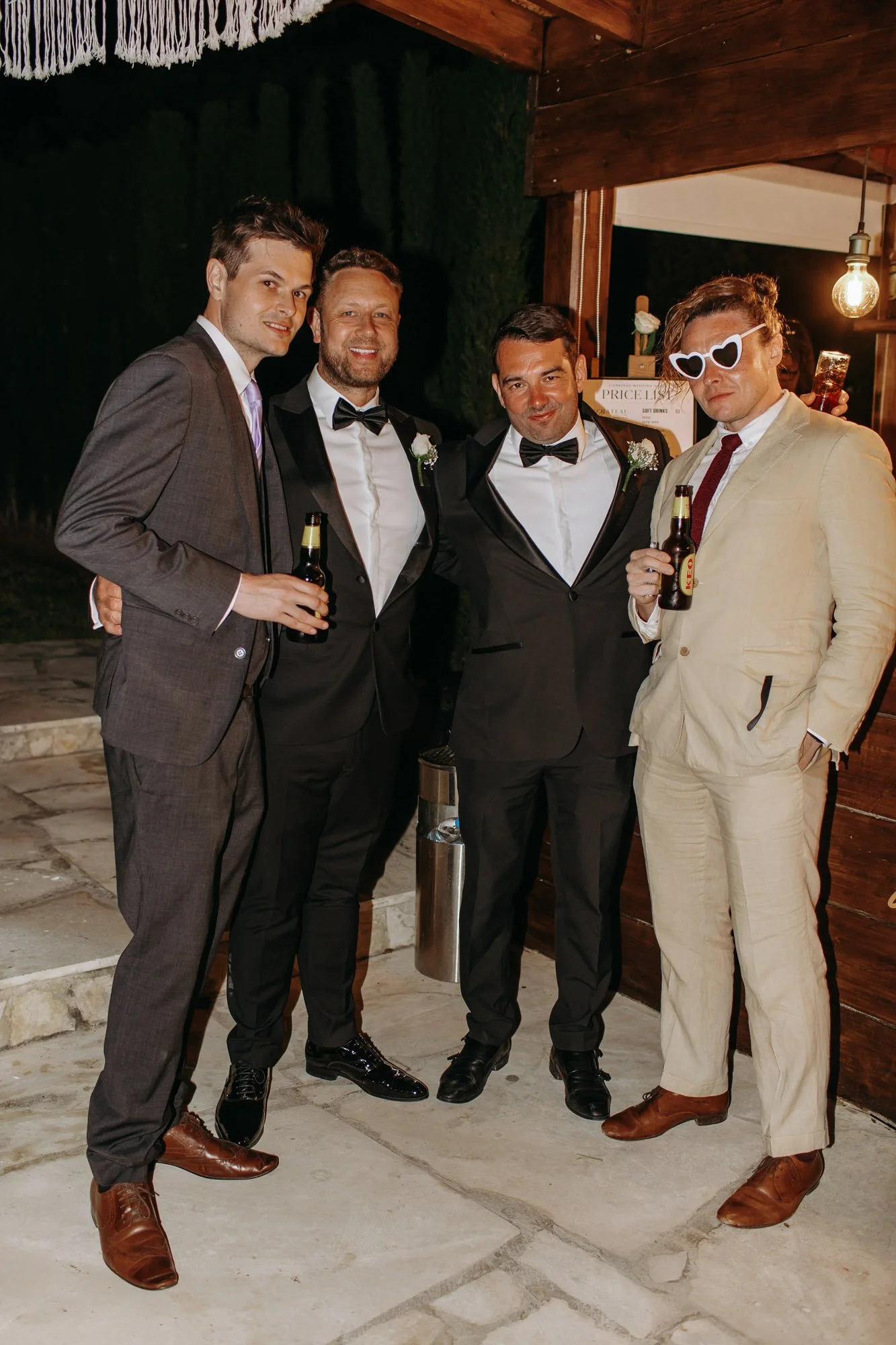 Group of five men dressed in formal suits and tuxedos, holding beer bottles, at a party or social event, with one wearing white heart-shaped sunglasses.