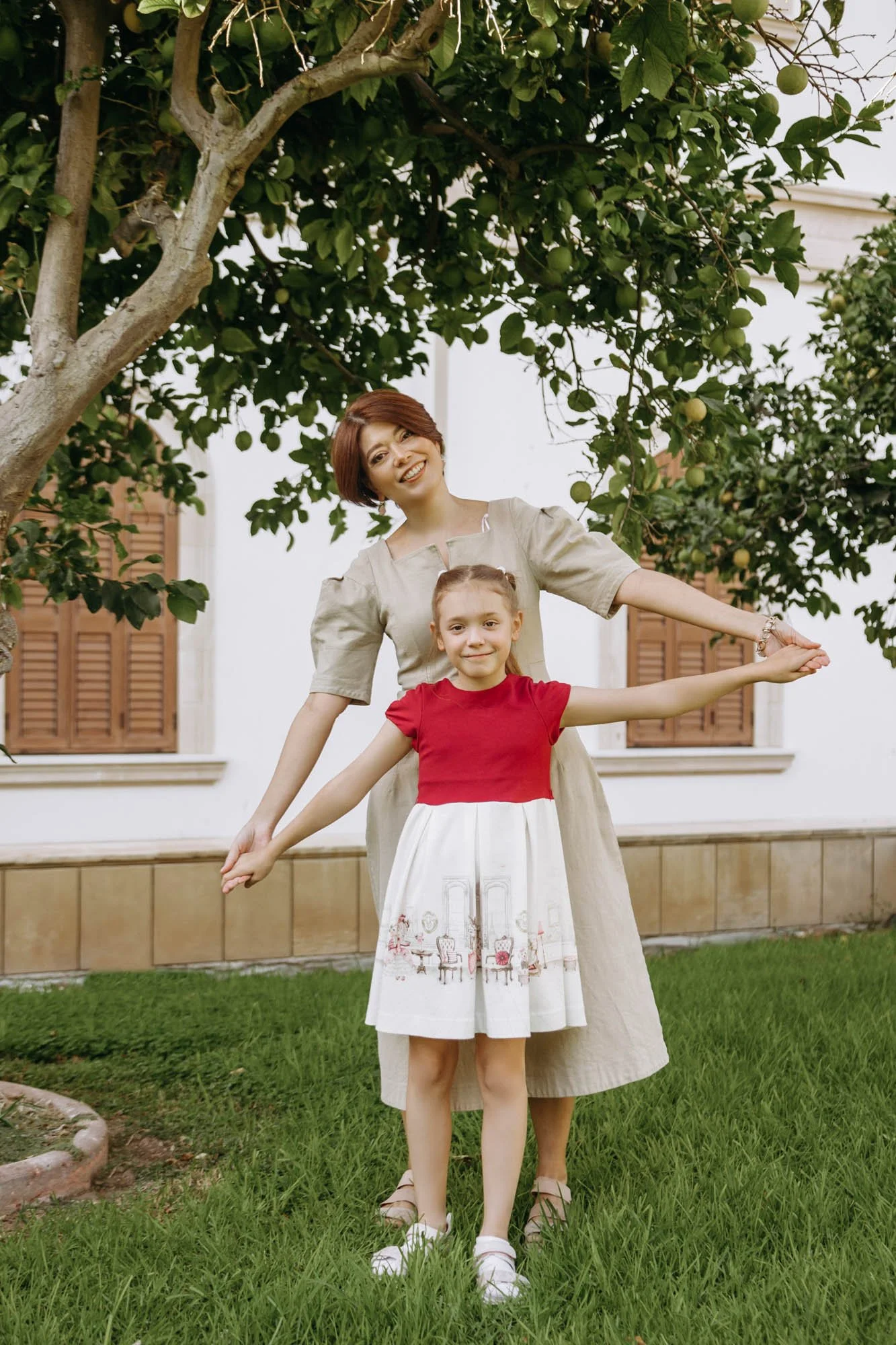 A woman and a young girl standing outside on grass near a tree, with her arms stretched out to the sides. The woman has short, brown hair and is smiling, wearing a beige dress. The girl has light brown hair tied in bun and is smiling, wearing a red t