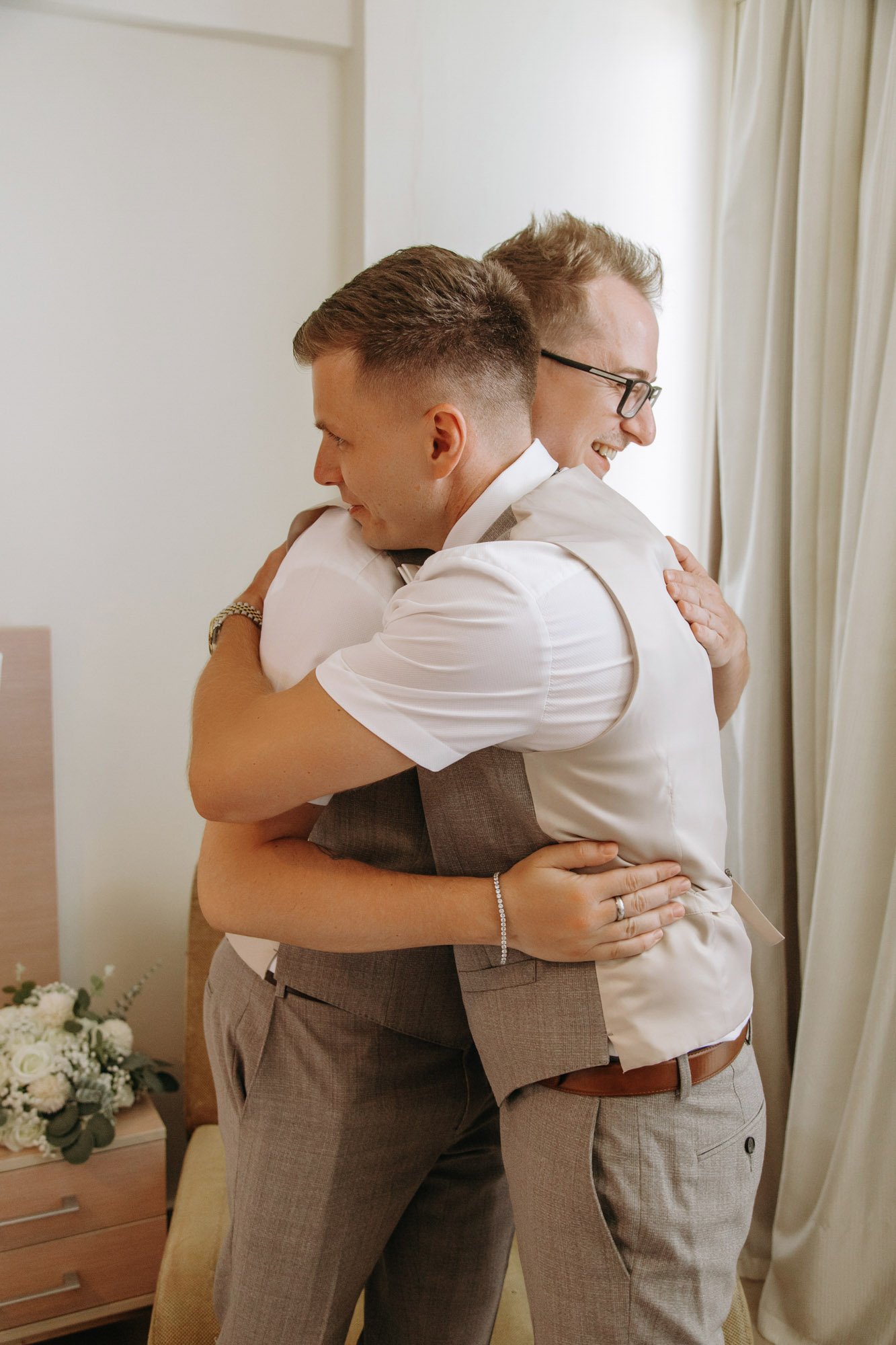 Two men sharing a warm embrace in a well-lit room, smiling with eyes closed, apparent happiness and affection.