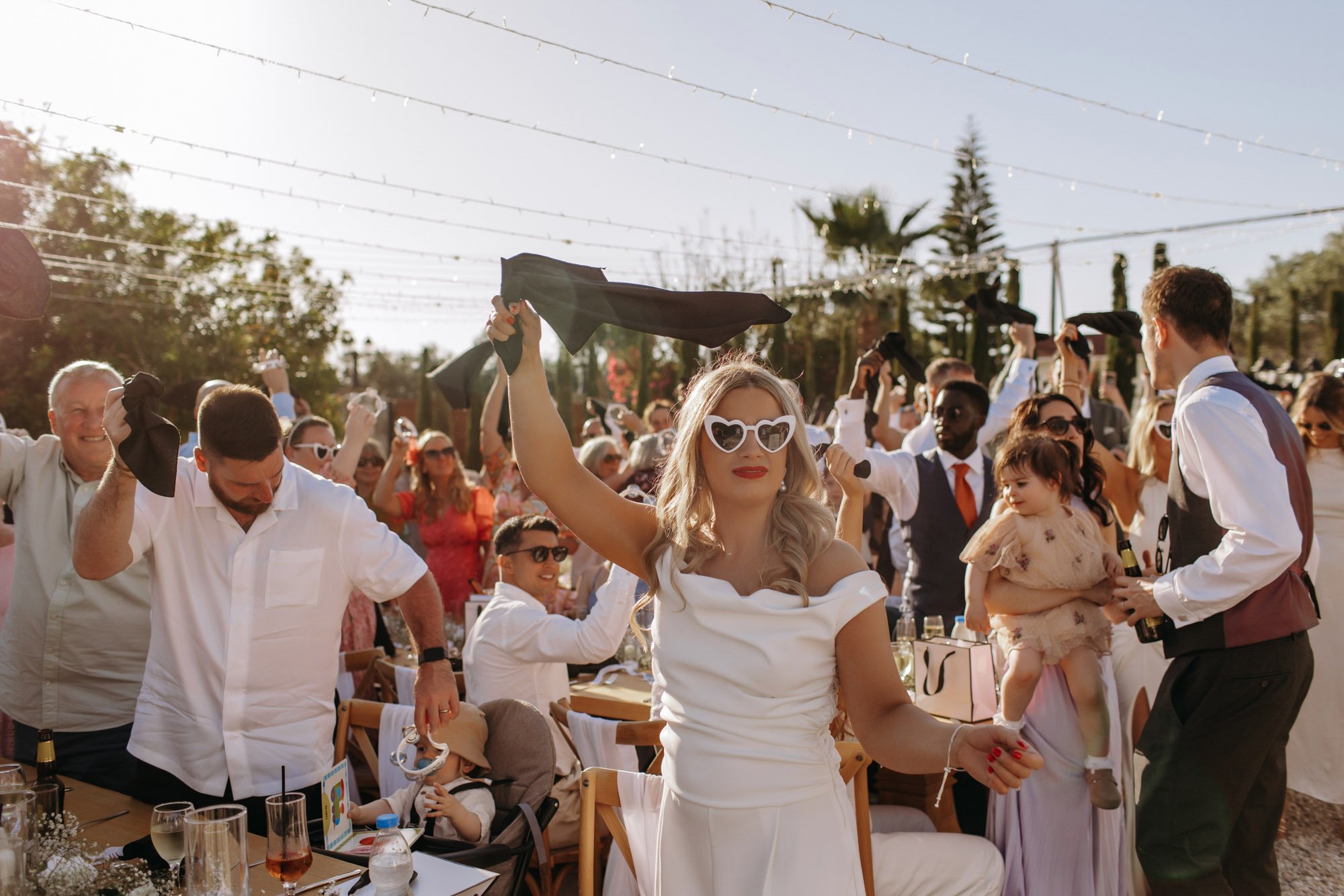 A woman in a white dress and heart-shaped sunglasses dancing at an outdoor celebration with many people in the background, some dancing and some sitting at tables.