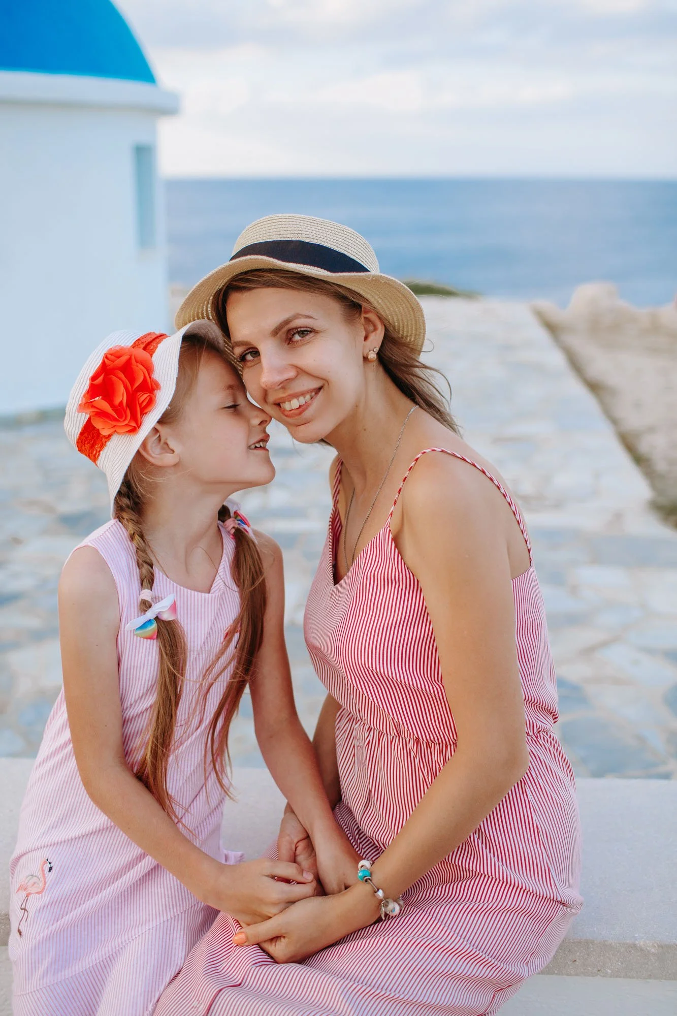 A woman and a young girl share a loving moment at the beach, holding hands and smiling. They are dressed in matching pink and white striped dresses and wearing summer hats. The background features the sea, sky, and a white building with a blue roof.