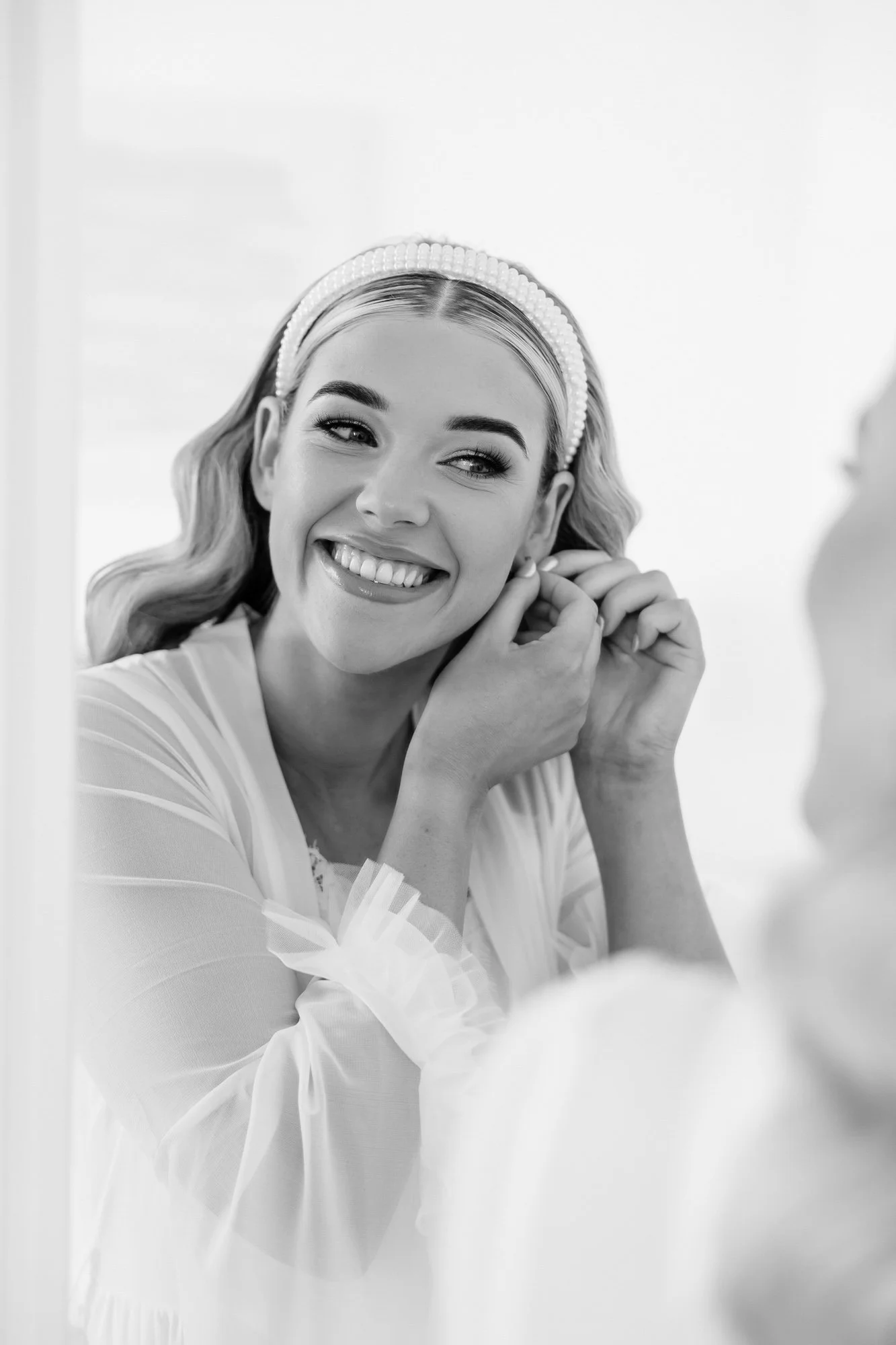 A woman smiling as she puts on earrings, looking in a mirror, wearing a pearl headband and a light-colored robe.