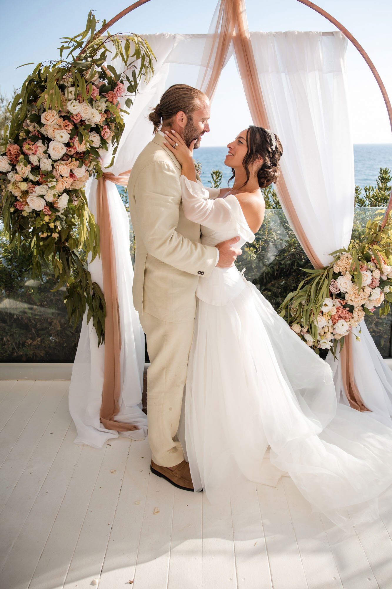 A bride and groom sharing a moment during their wedding ceremony with a backdrop of the ocean and sky, decorated with white and pink flowers and draped fabric.