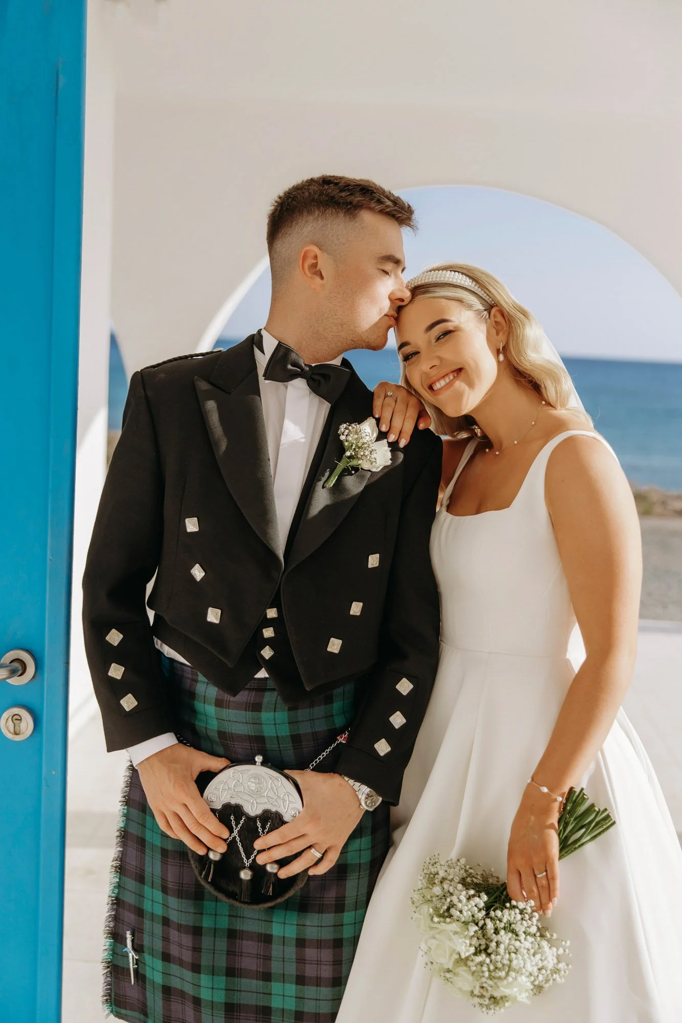 A bride and groom with the groom kissing the bride on her forehead as she smiles. The bride wears a white dress and holds a bouquet, while the groom is dressed in a traditional black tuxedo with a tartan kilt and is holding a small bag.
