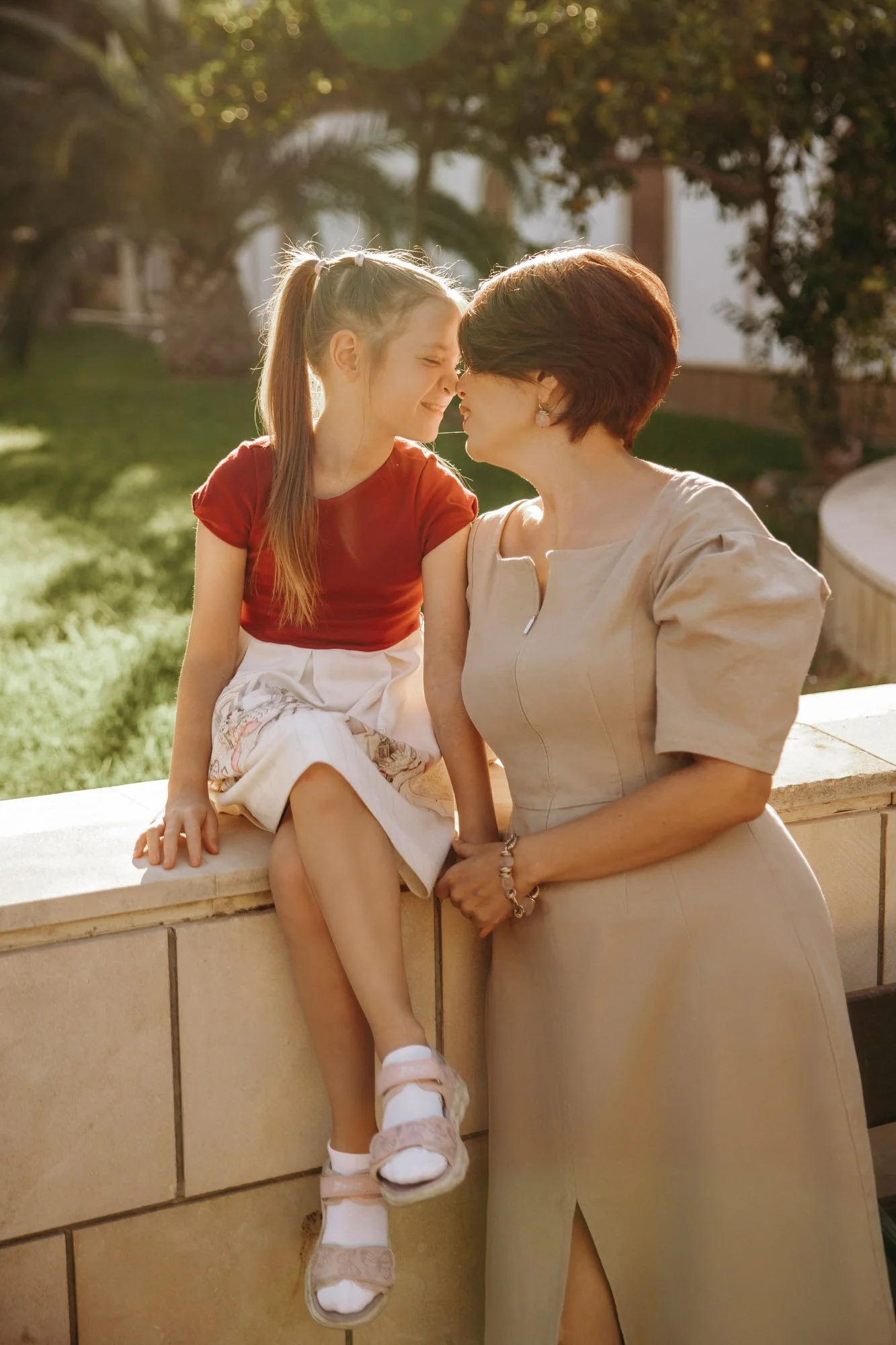 A young girl and an older woman are close together, smiling, with their foreheads touching outdoors during daytime. The girl is sitting on a ledge wearing a red top and white skirt, and the woman is standing beside her wearing a beige dress. Sunlight