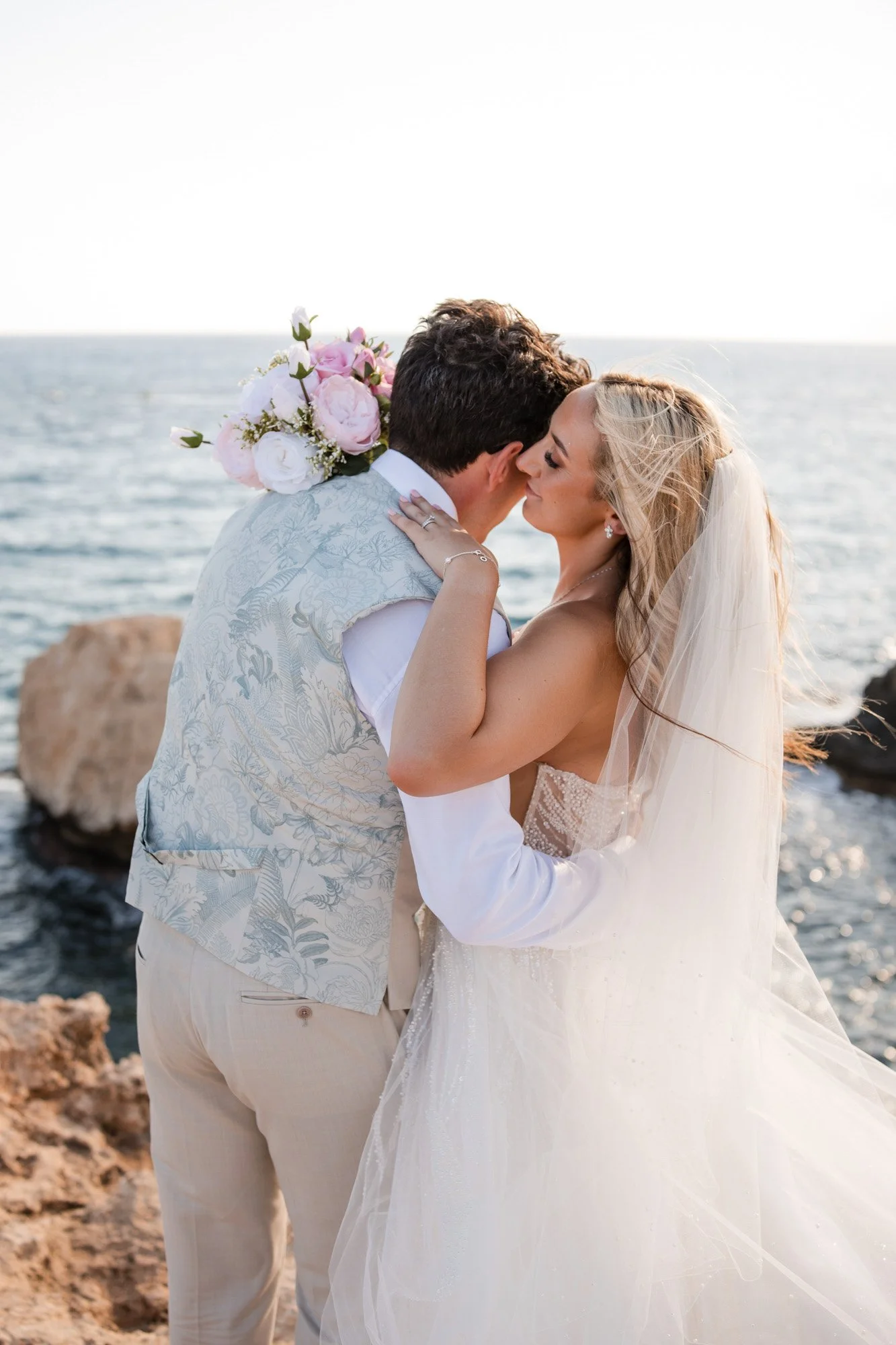 A bride and groom kissing on a rocky shore during sunset, with the ocean in the background.