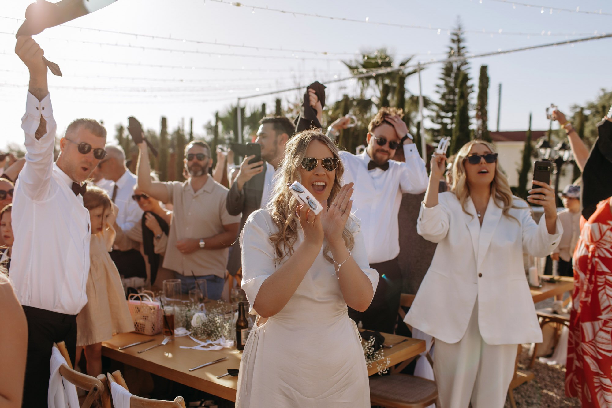 Group of people celebrating outdoors, some wearing sunglasses, holding phones and drinks, with a woman in white at the center smiling and clapping, in a sunny setting with string lights and trees in the background.
