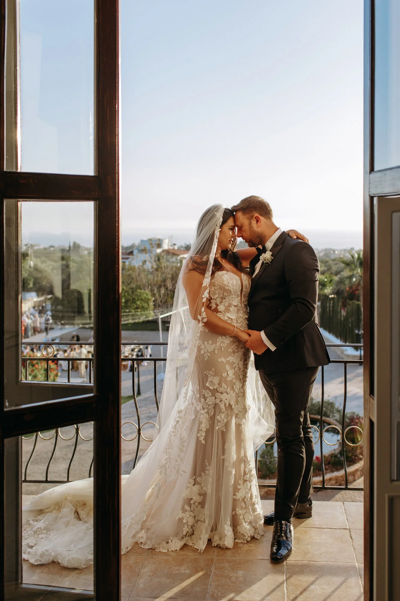 Bride and groom sharing an intimate moment on their wedding day, holding hands, with the bride in a lace wedding gown and veil, and the groom in a black suit, standing on a balcony at sunset.