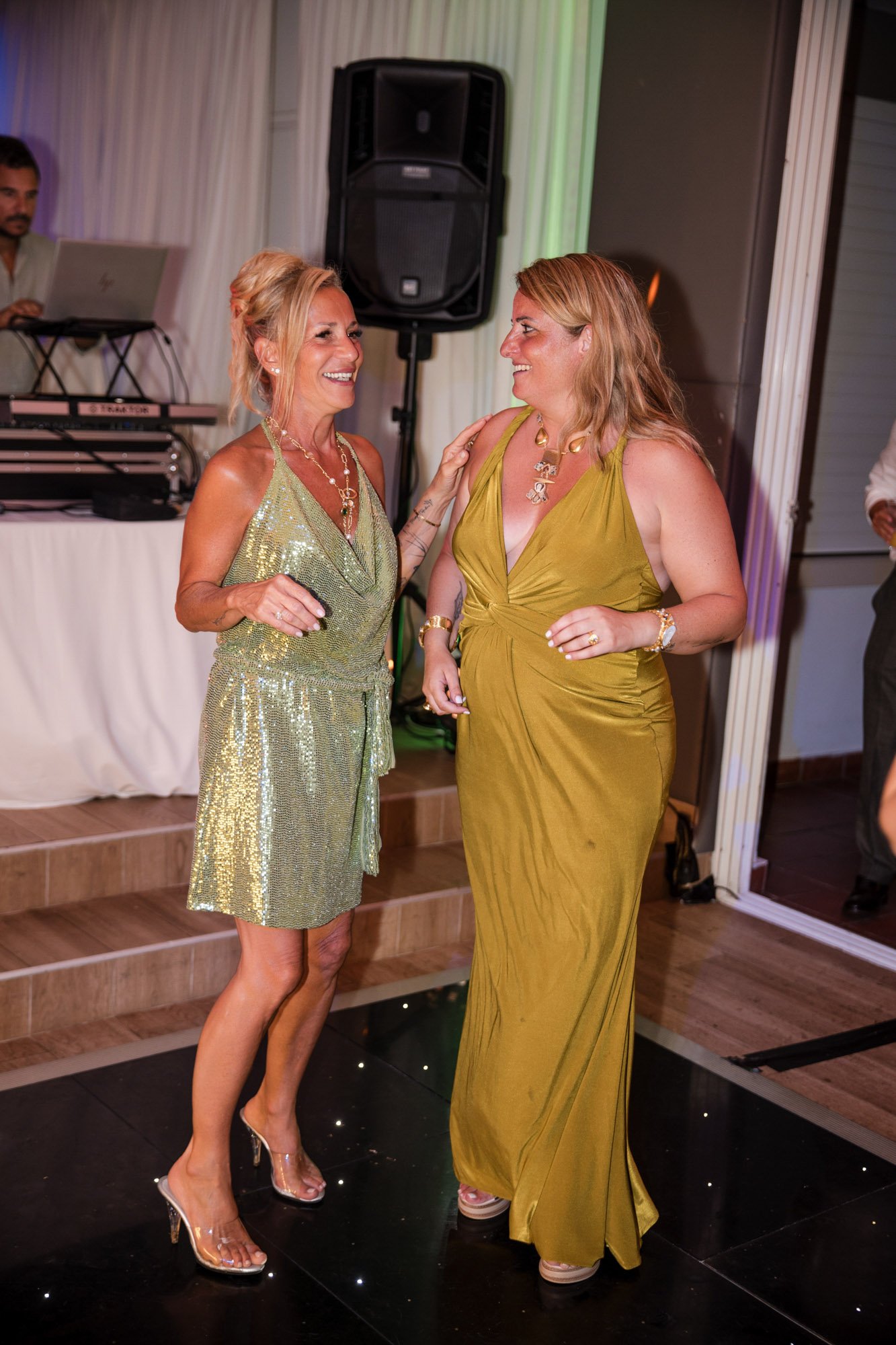 Two women in elegant dresses smiling and dancing at a party, with a DJ in the background.