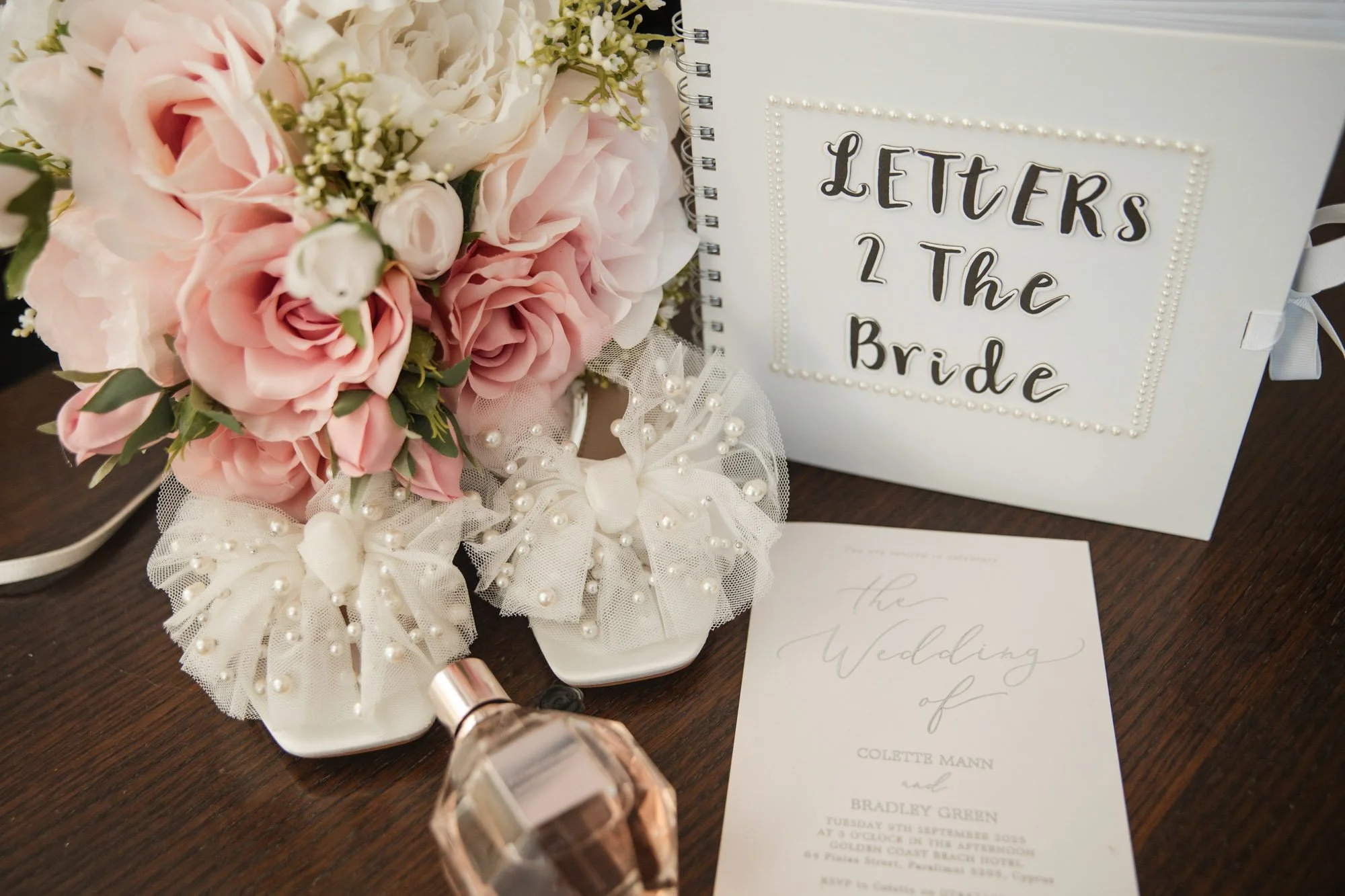 A wedding table centerpiece with pink roses, white flowers, and decorative white tulle bows with pearls, alongside a wedding invitation, a perfume bottle, and a white book labeled 'Letters to the Bride'