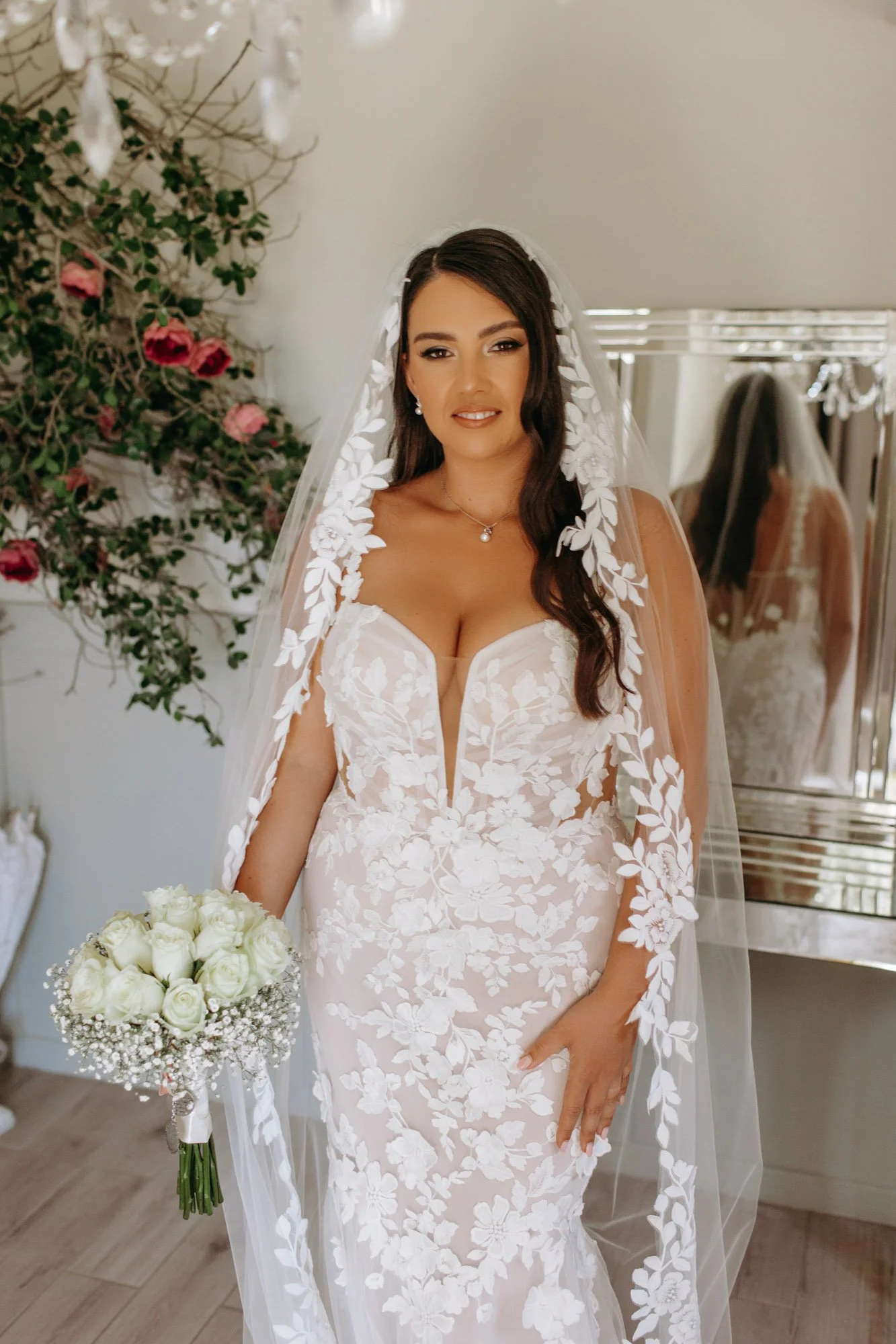 Bride with dark hair in a white floral lace wedding gown holding a bouquet of white roses, standing indoors near a mirror and floral arrangement.