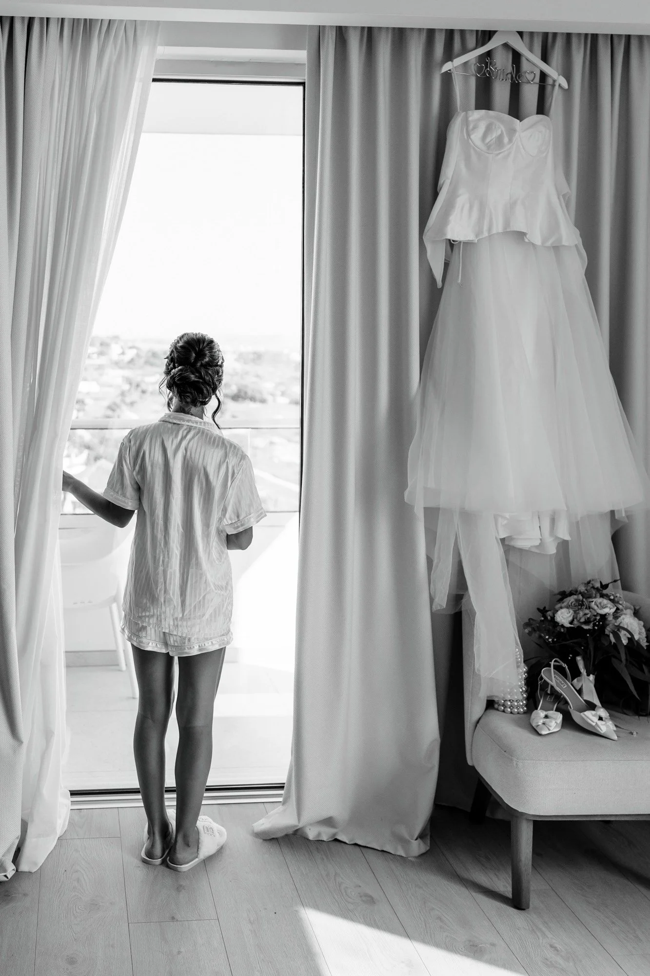 A woman in pajamas standing by a large window, looking outside; a wedding dress hanging on a hook on the curtain rod, with wedding shoes and flowers on a bench nearby in a room.