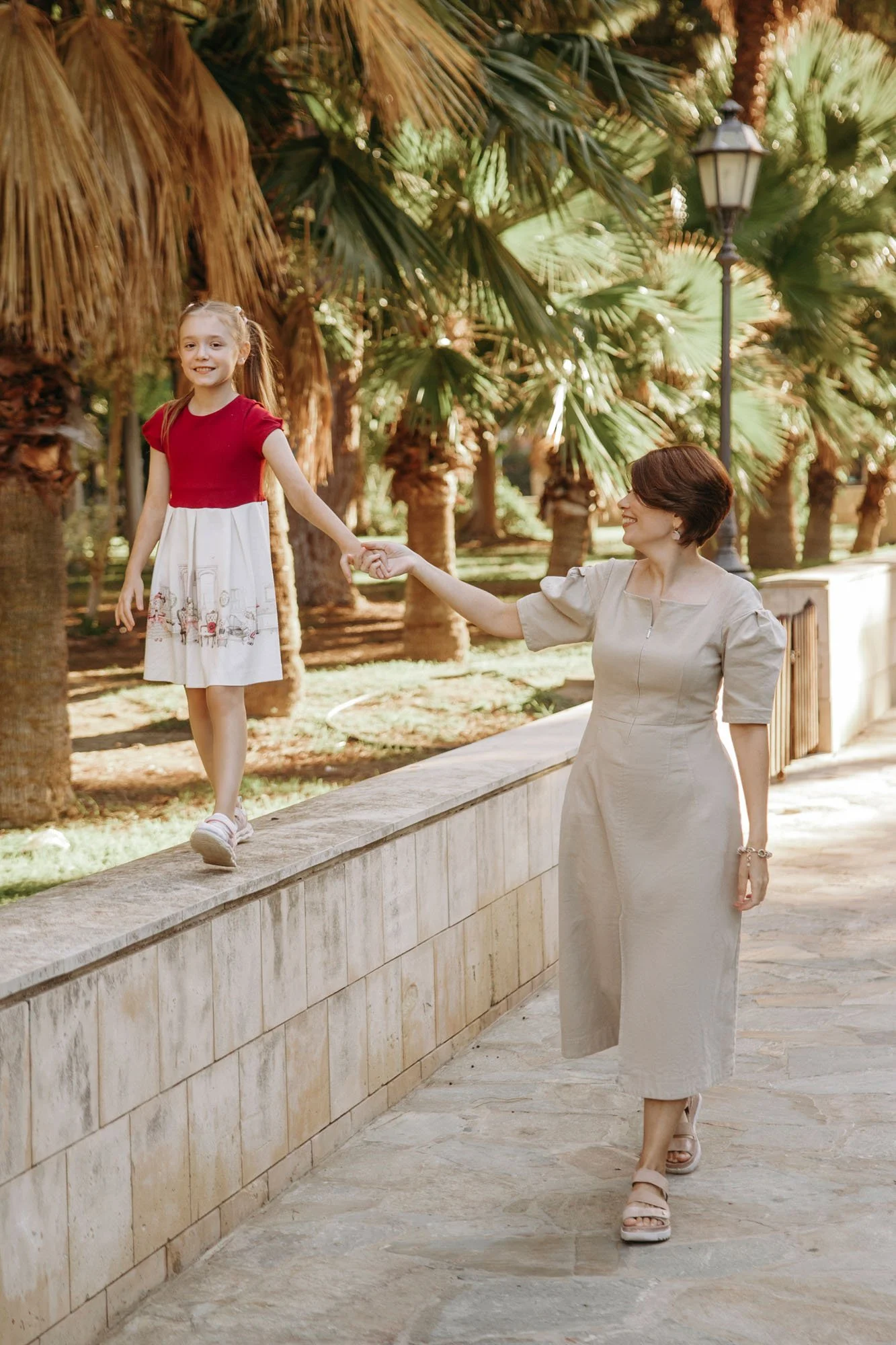 A woman and a girl holding hands; the girl is walking on a low wall and the woman is walking beside it, surrounded by palm trees and outdoor lighting.