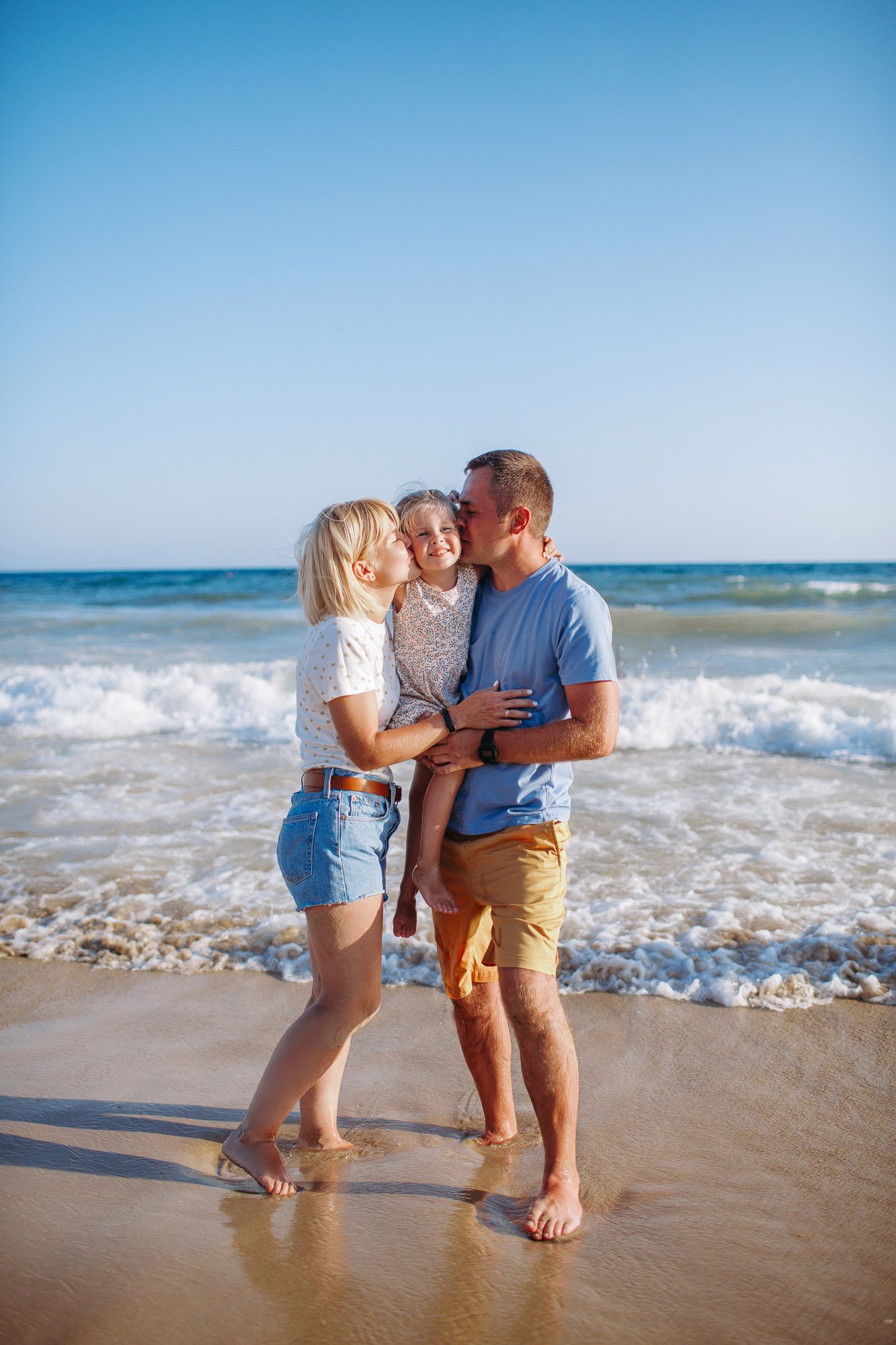 A family of three, two women and a man, standing on the beach with the ocean waves in the background. The women are kissing the young girl on her cheeks as the man holds her. The group appears to be enjoying a sunny day together.