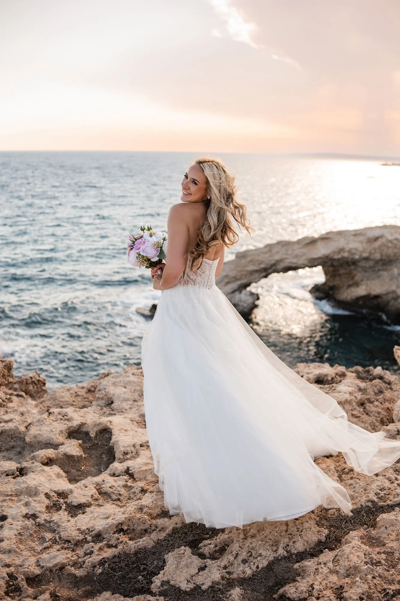 Bride in a white wedding dress holding a bouquet, standing on rocky coast with ocean and sunset in background, smiling over her shoulder.