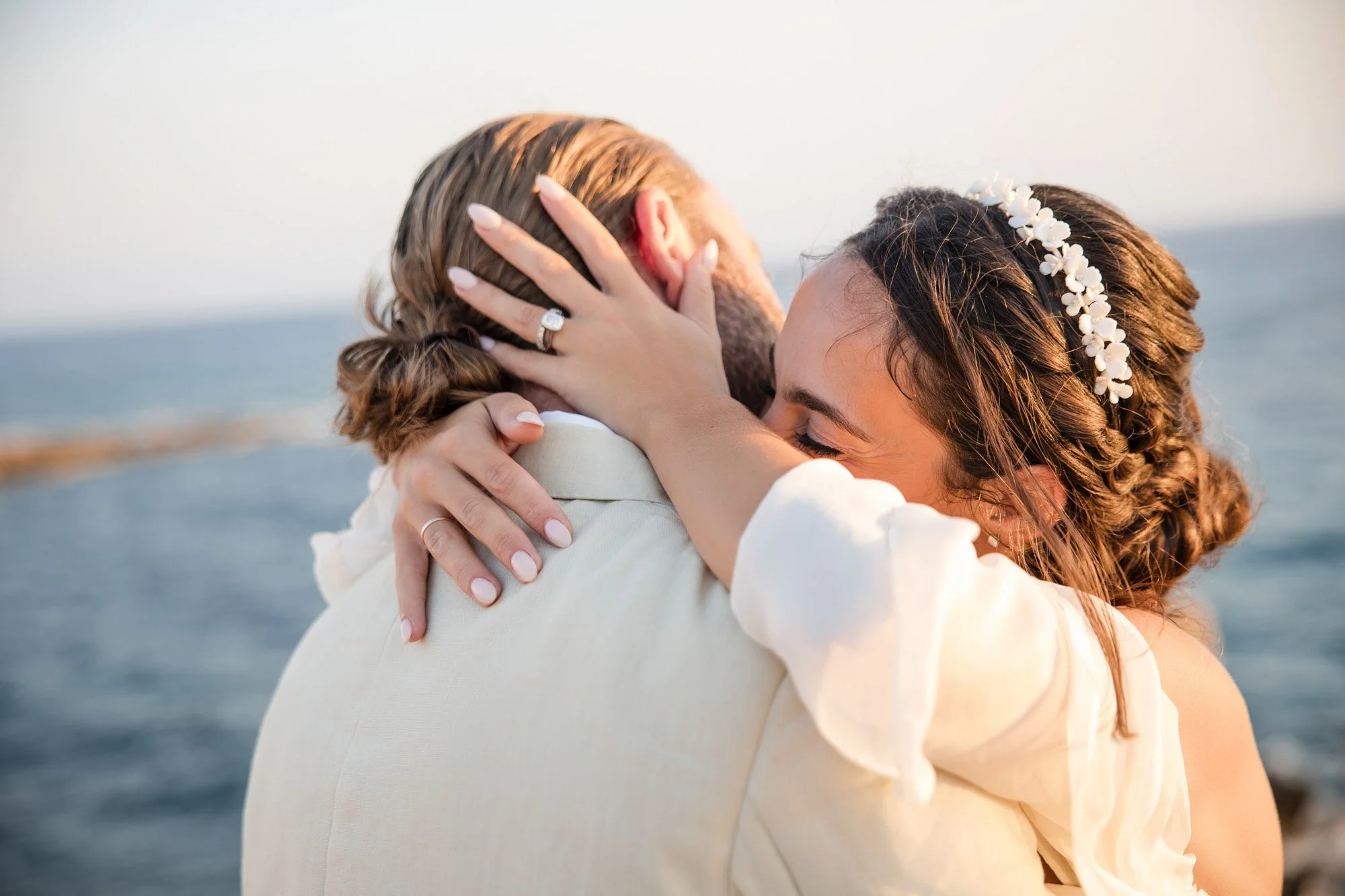 A woman with a floral headband embracing a man by the water, with her eyes closed and a joyful expression.