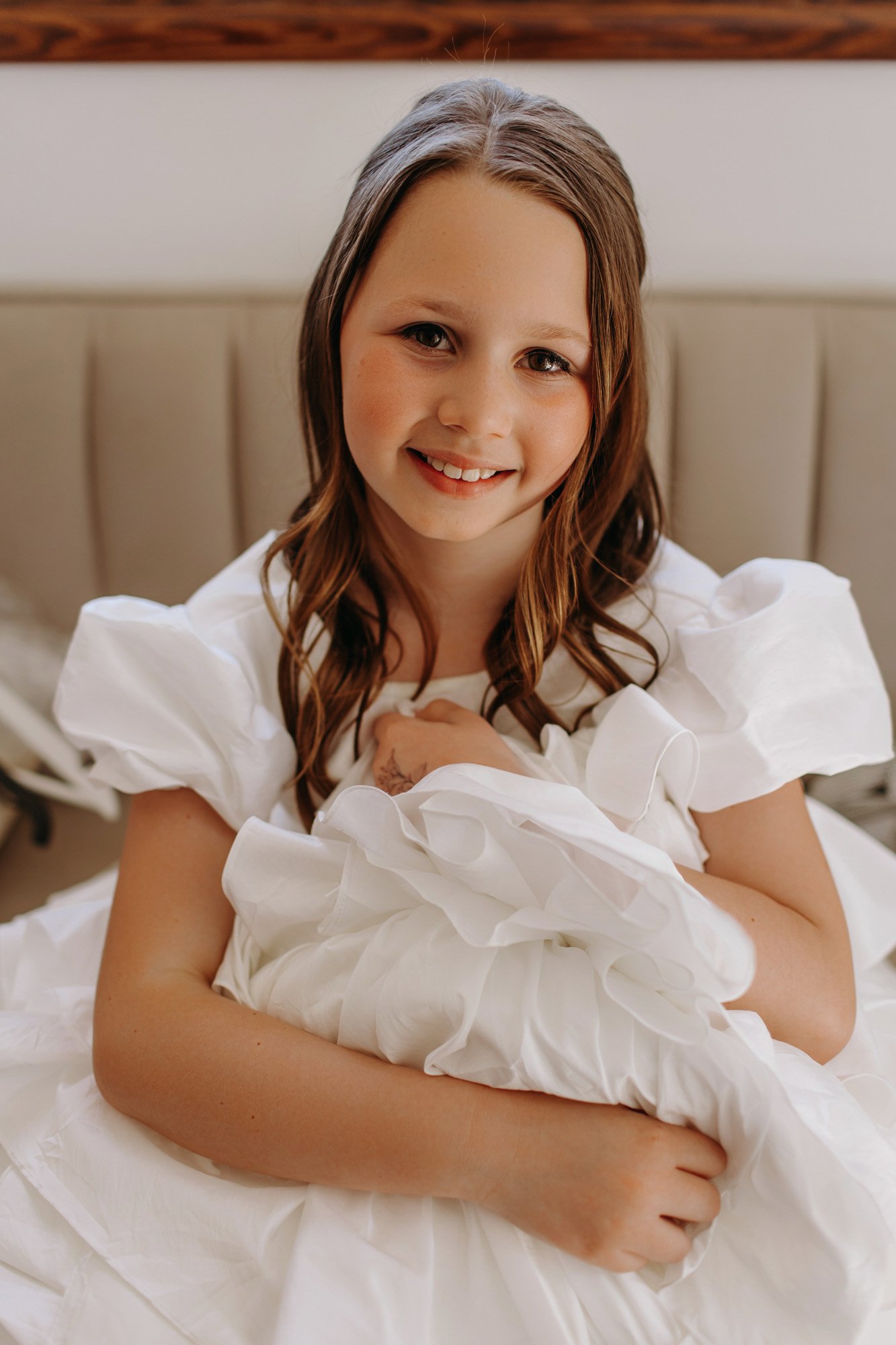 A young girl smiling while sitting on a bed in a white dress with puffed sleeves, holding a white fluffy blanket.