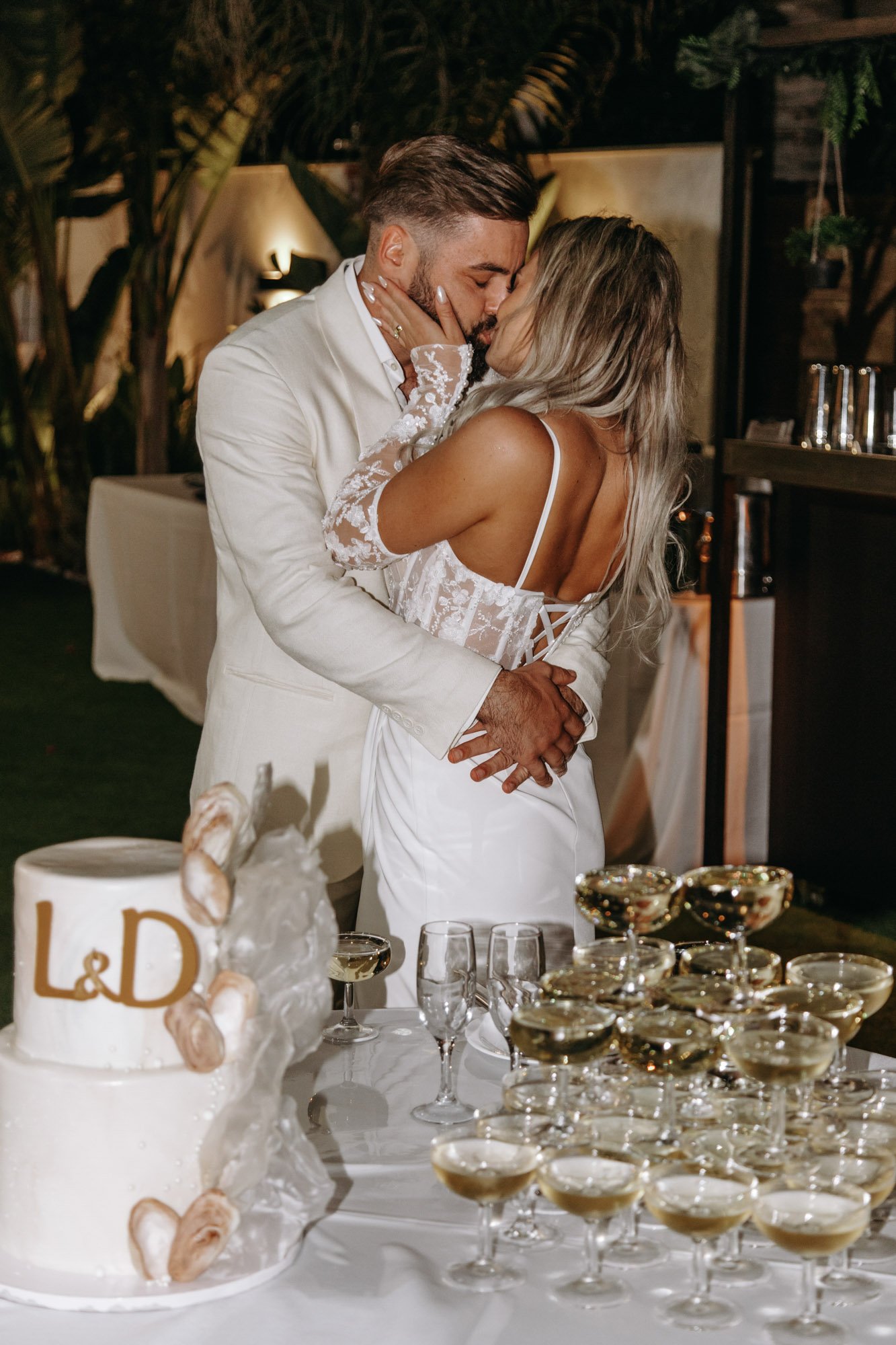 A couple kissing at their wedding reception, with a cake and champagne glasses on the table in front of them.