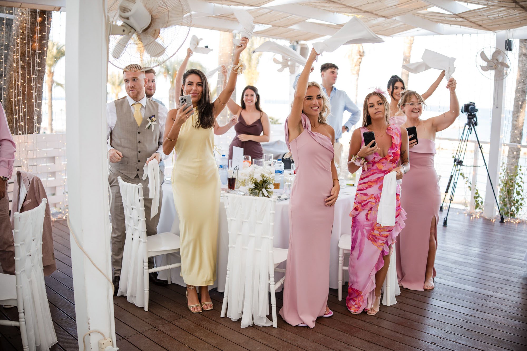 Group of people at a wedding reception, standing and raising napkins in the air, some taking photos or videos with their phones, indoors with decorated setting and tableware.
