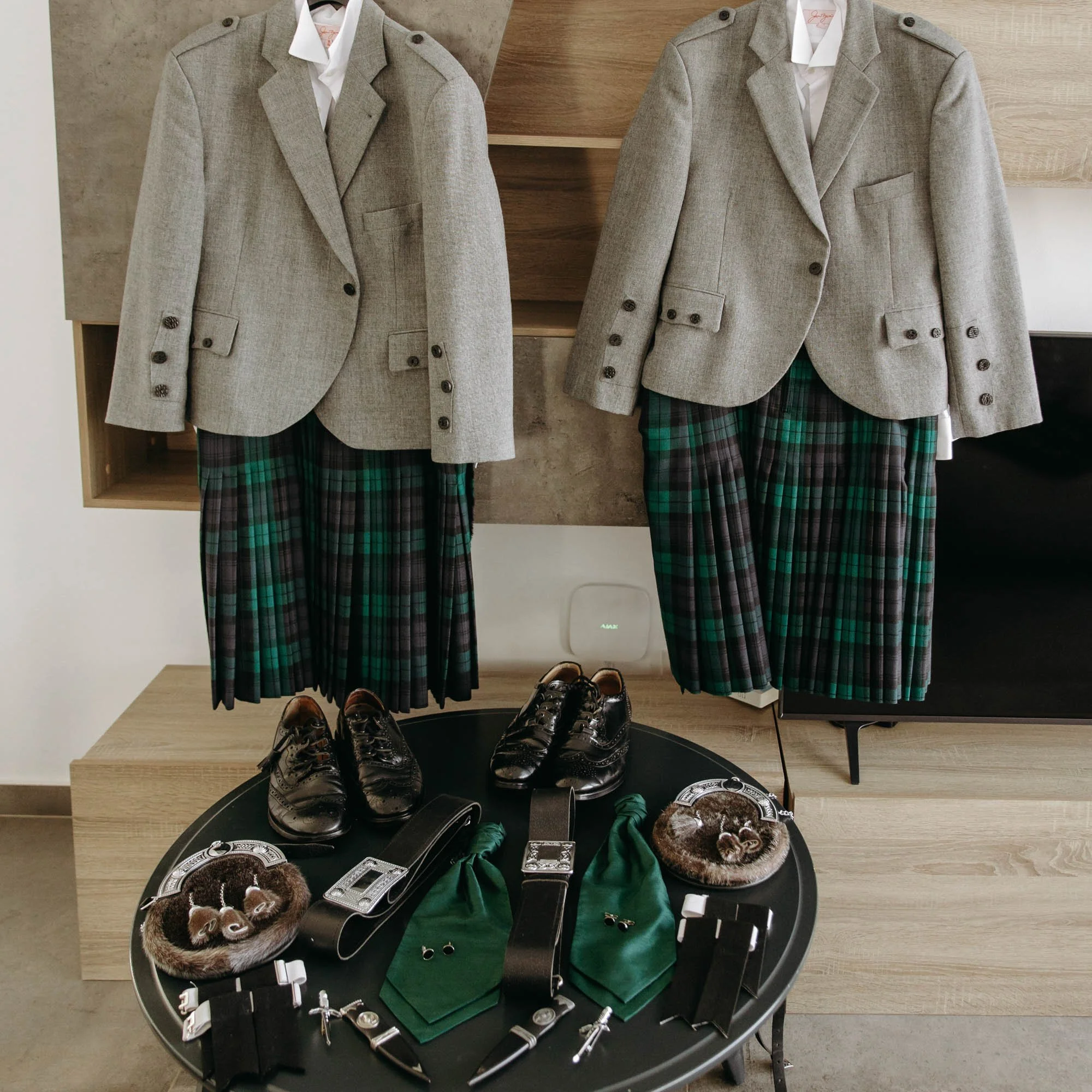 Two outfits featuring gray blazers, green and black plaid skirts, and white shirts hanging on a wall. A variety of shoes, handbags, and accessories are arranged on a black round table in front of the outfits.