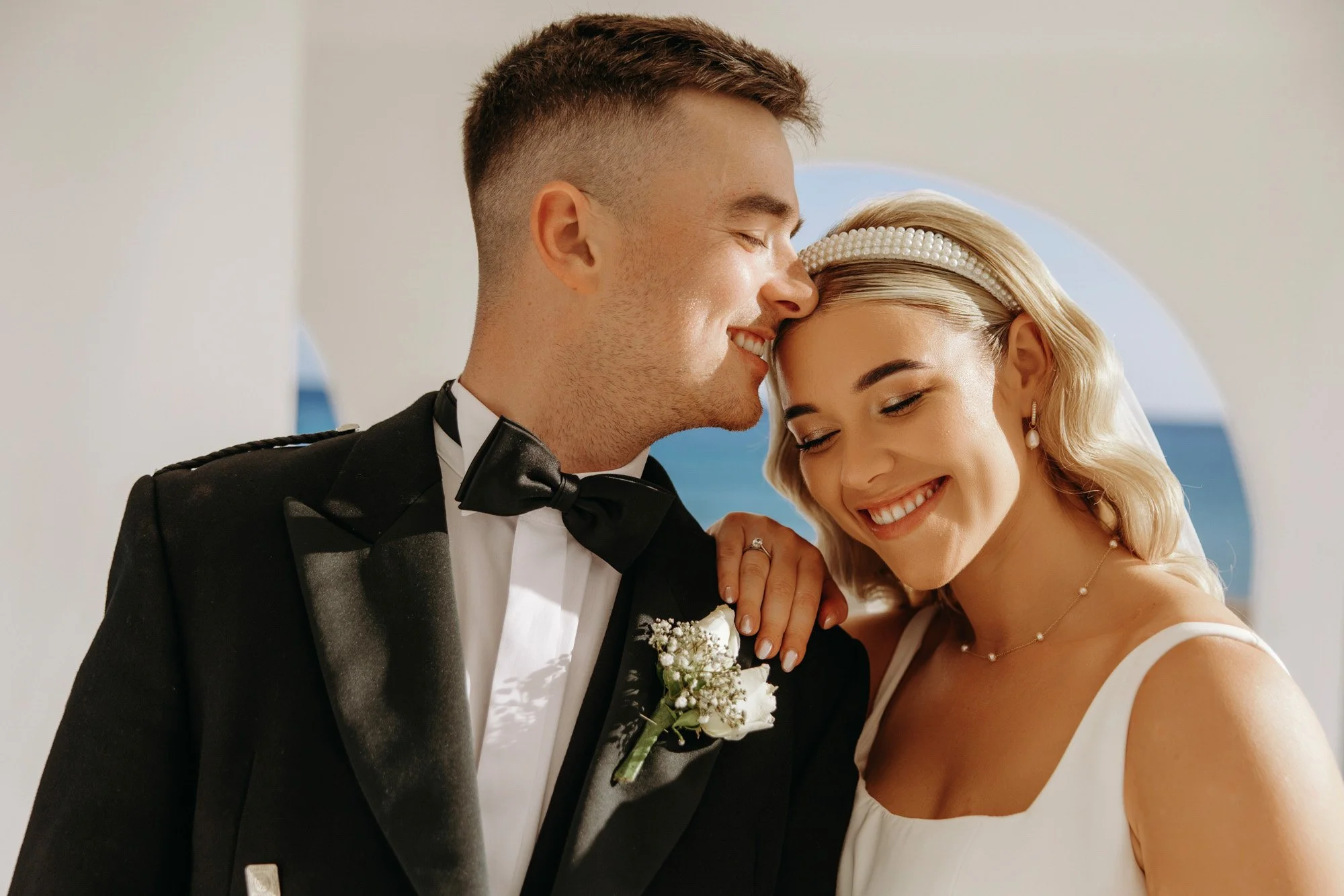 A happy bride and groom on their wedding day, smiling with their faces close together, in front of a scenic ocean view.