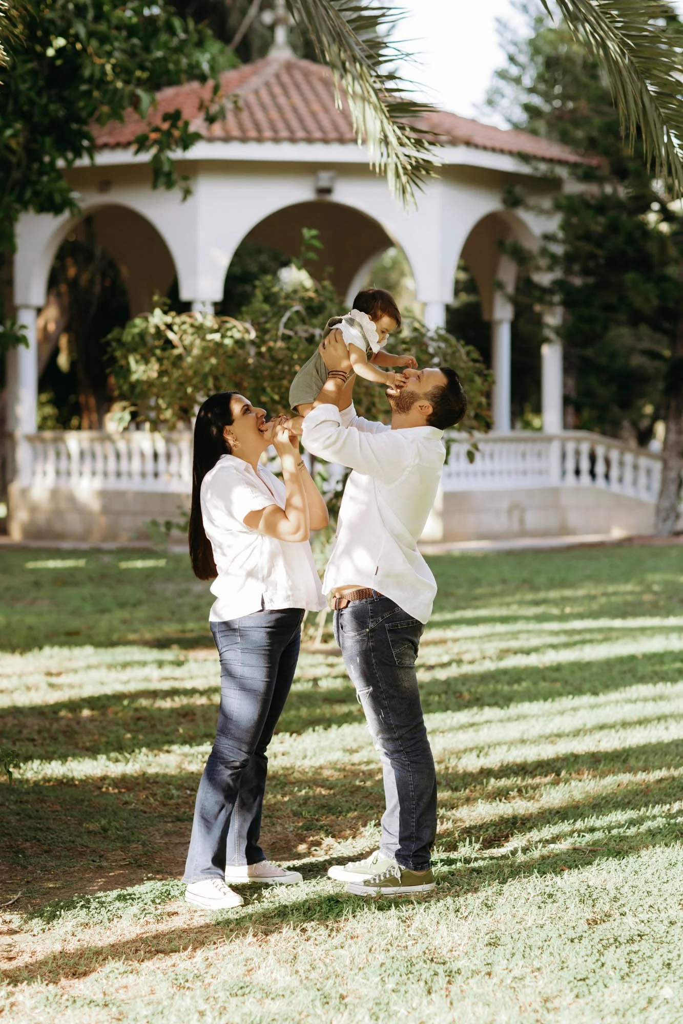 A family of three outdoors with a house in the background. The father lifts a young girl high in the air, and the mother stands beside them smiling, in a sunny, green setting.