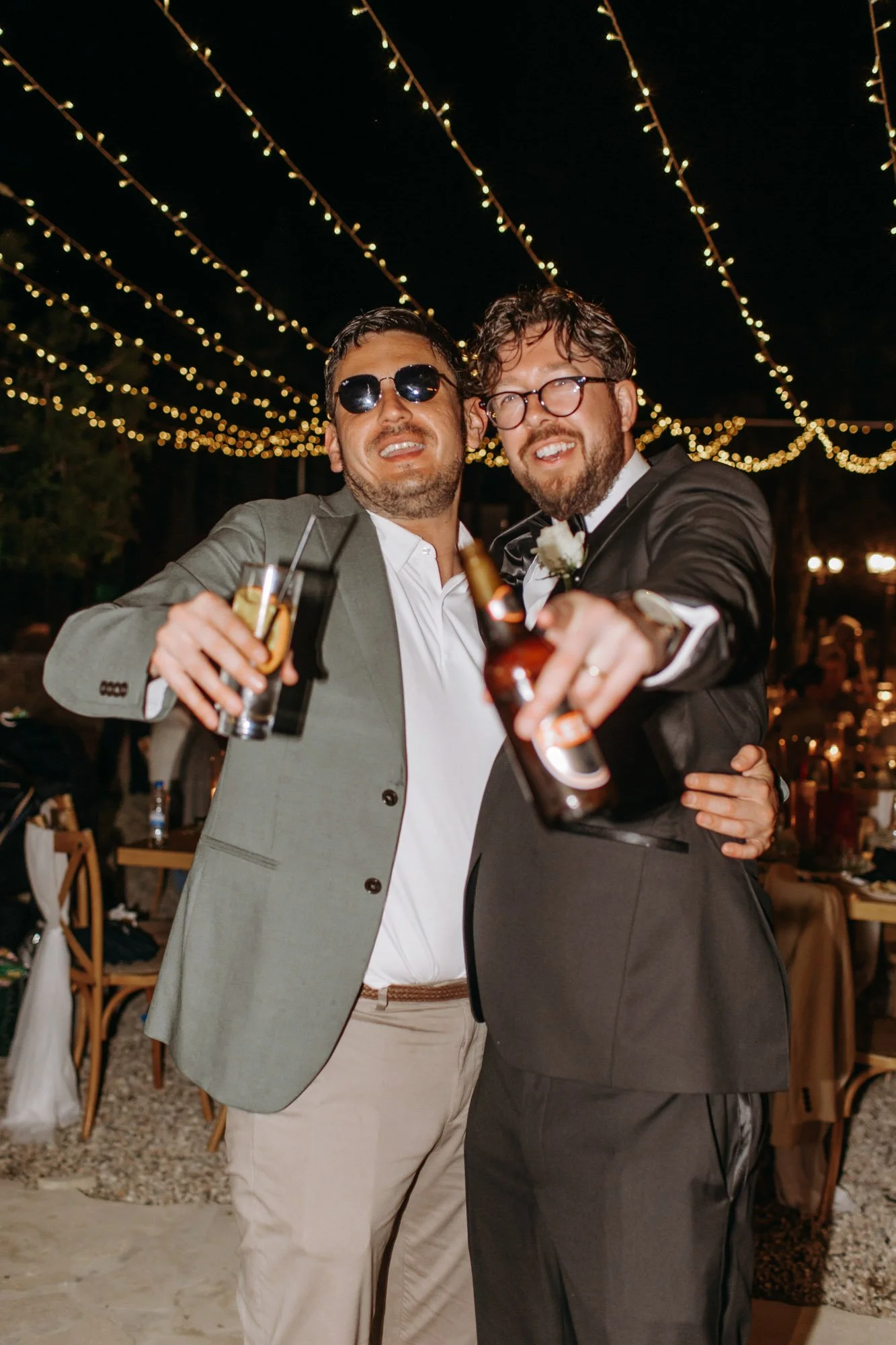 Two men dressed in suits celebrating at an evening outdoor event under string lights, holding drinks and smiling.