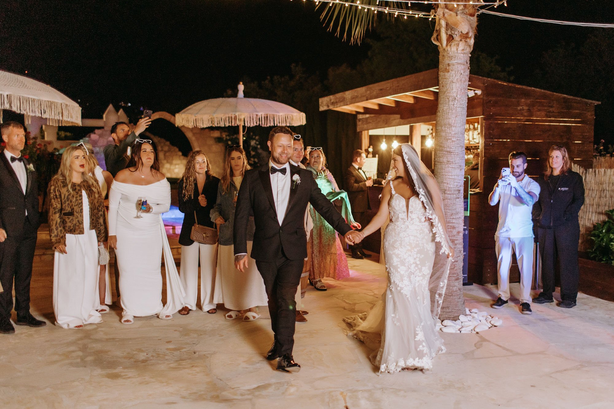 A newlywed couple, the groom in a black tuxedo and the bride in a white lace wedding gown, holding hands and walking on a dance floor at night. Guests are gathered around, some taking photos, under warm string lights in an outdoor wedding setting wit