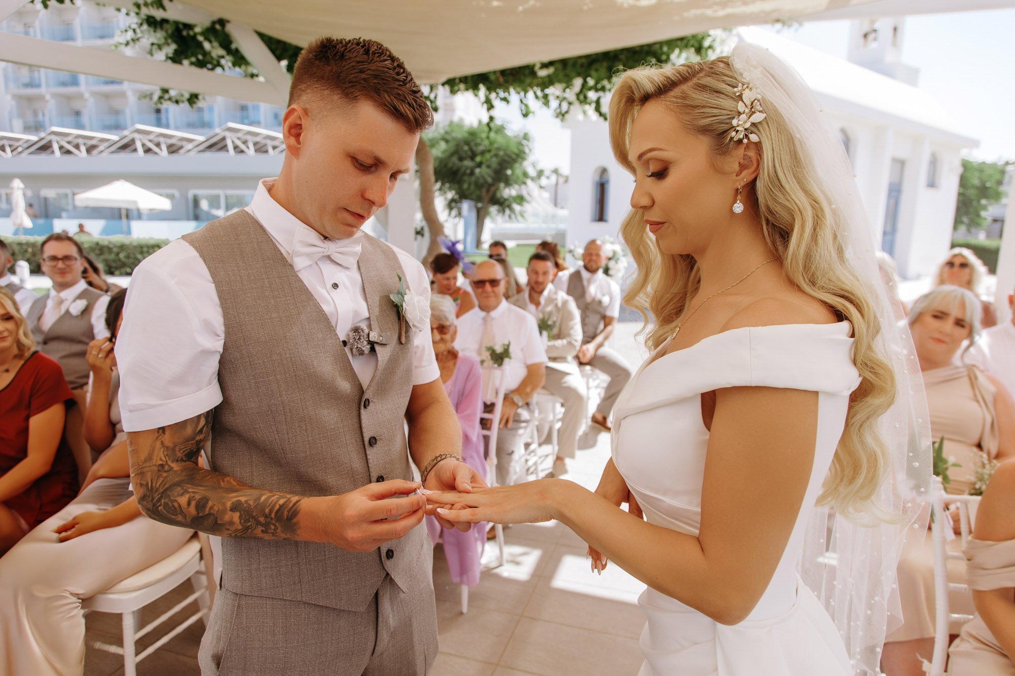 A bride and groom exchanging wedding rings during an outdoor wedding ceremony, surrounded by seated guests.