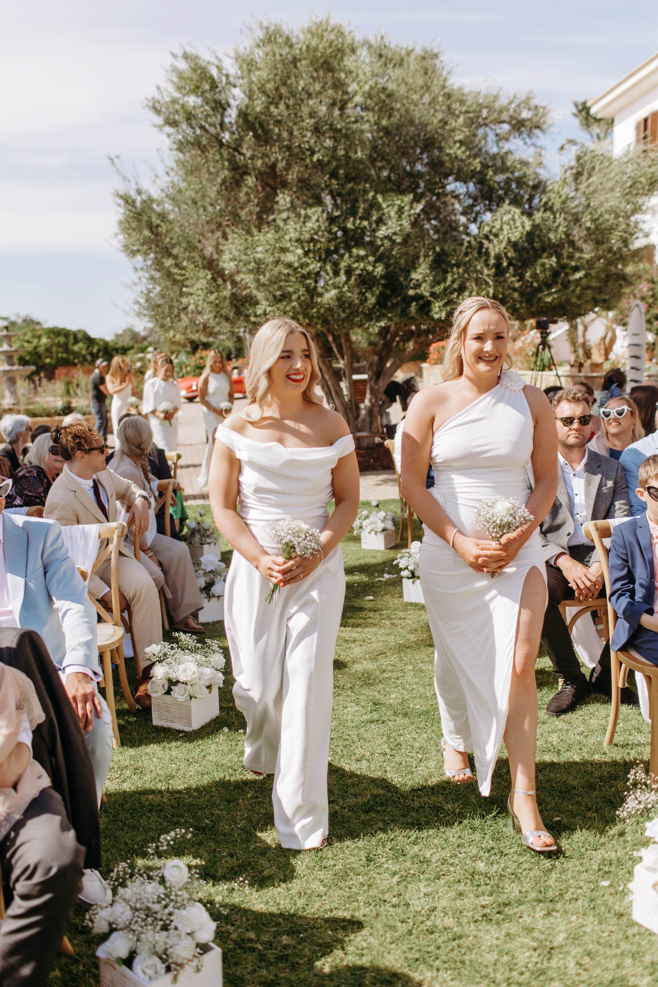 Two brides in white dresses walking down the aisle at an outdoor wedding ceremony surrounded by seated guests, with a large tree and clear skies in the background.