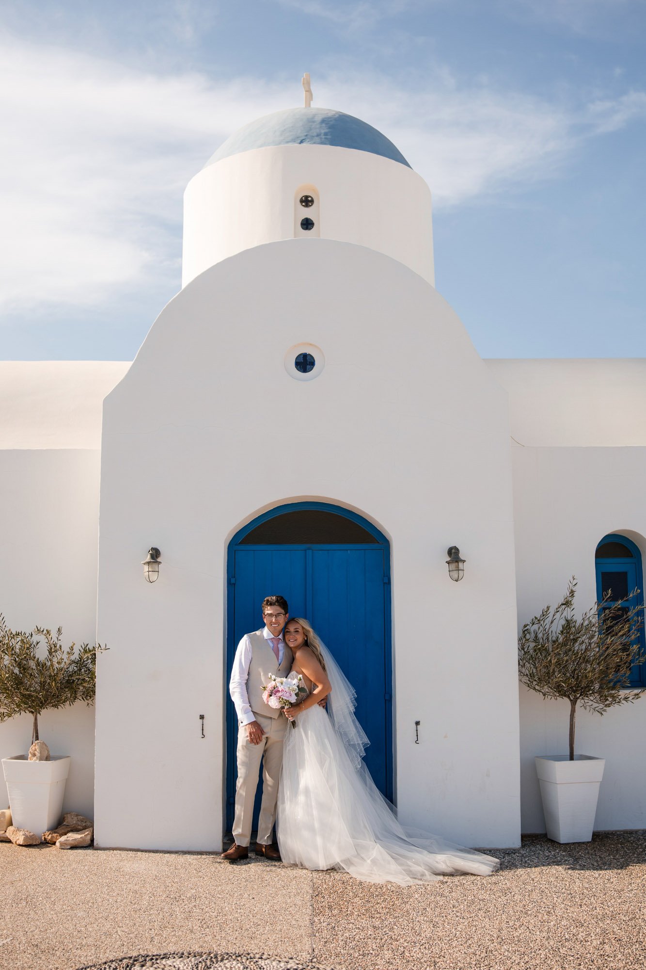 A newlywed couple standing in front of a white and blue church, with the bride holding a bouquet of flowers.