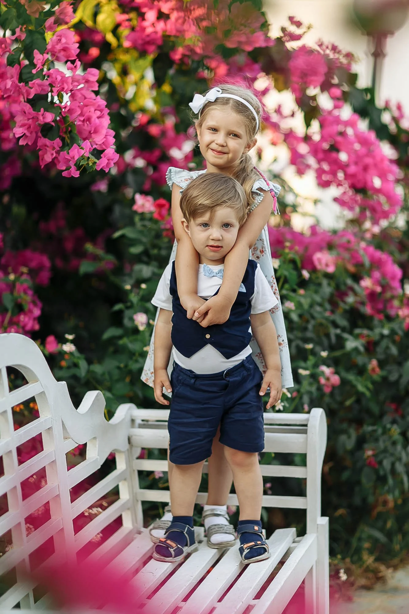 Two young children, a girl with a white bow in her hair and a boy, standing on a white garden bench in front of pink flowers, with a white fence nearby.