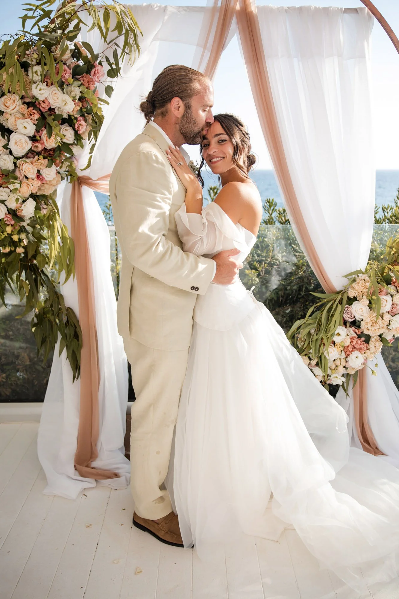 Bride and groom embrace during their wedding ceremony near a floral arch overlooking the ocean.